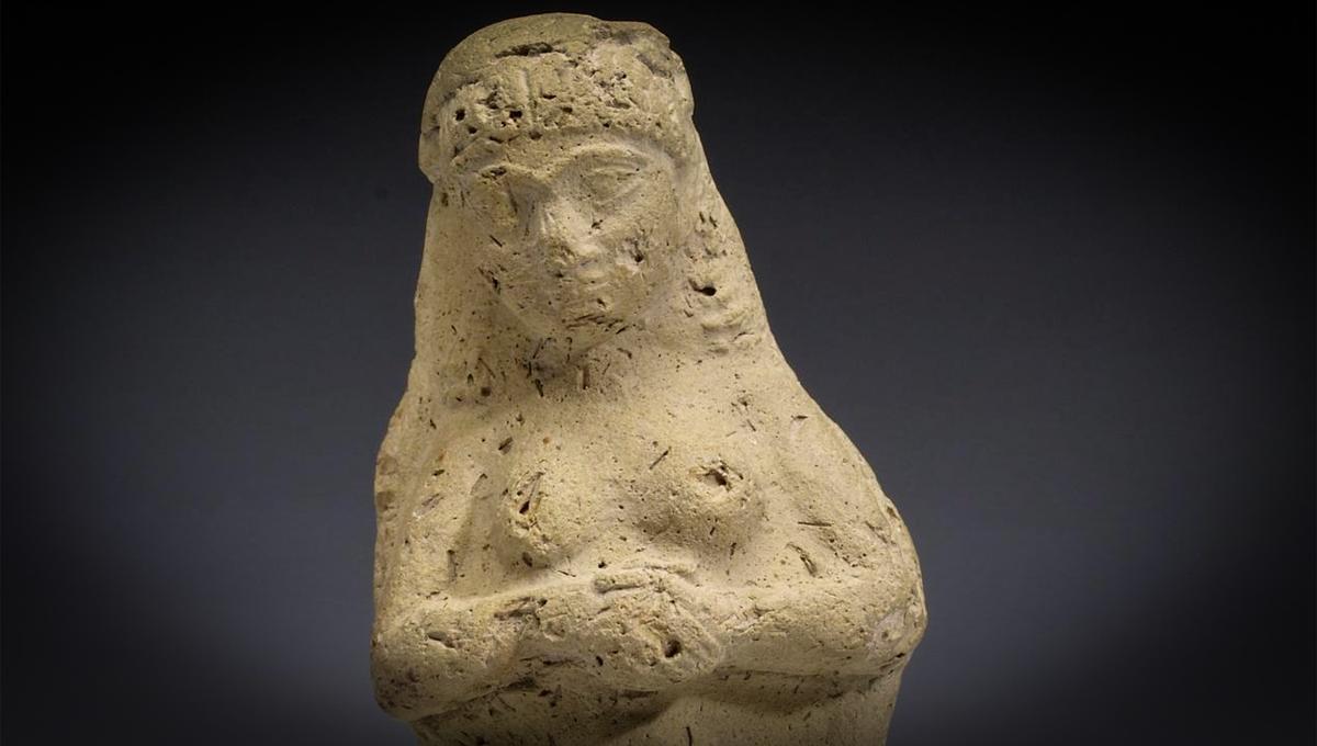 Clay sculpture of a standing female figure with folded arms against a dark background, worn and aged with visible markings