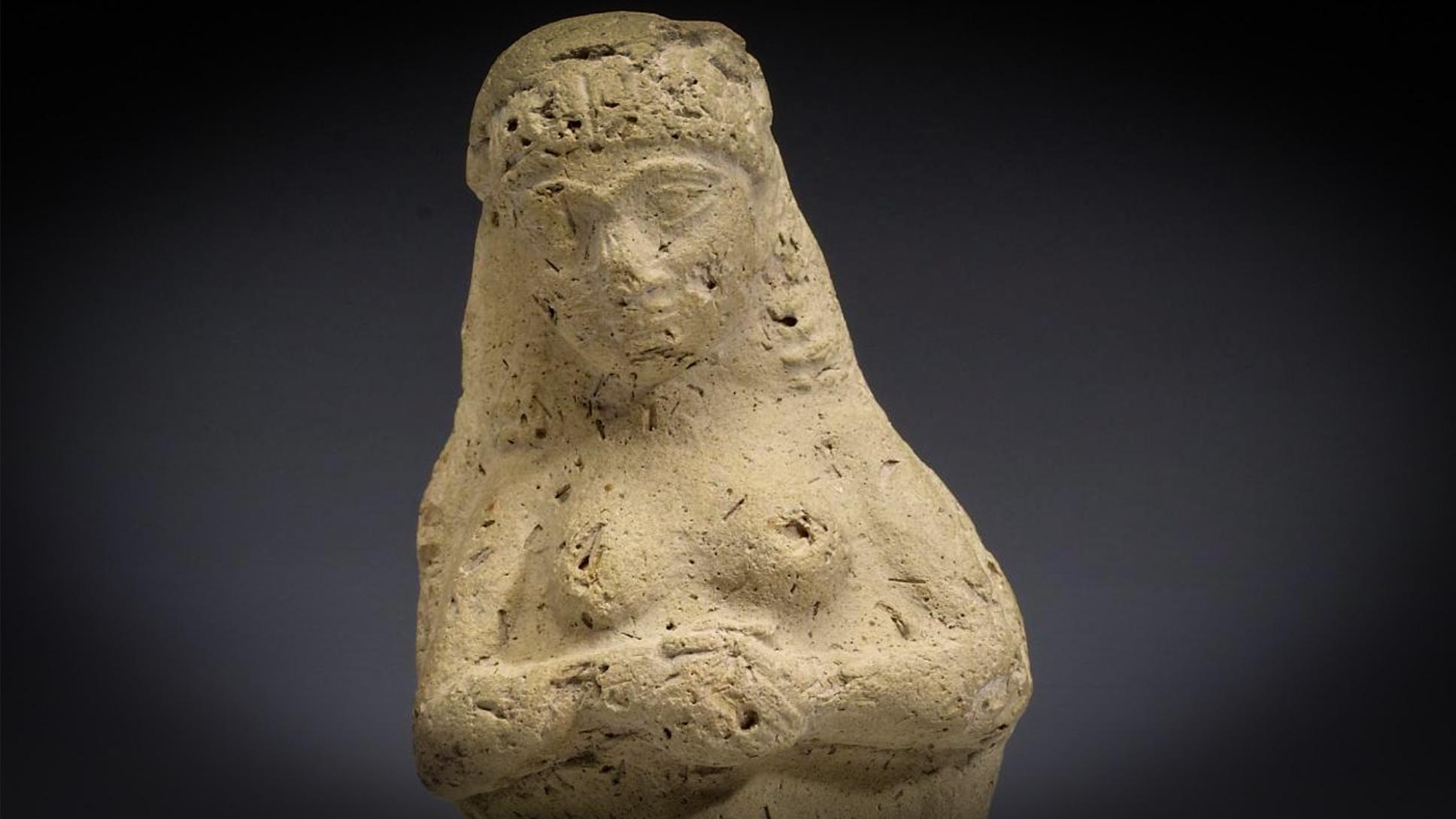 Clay sculpture of a standing female figure with folded arms against a dark background, worn and aged with visible markings