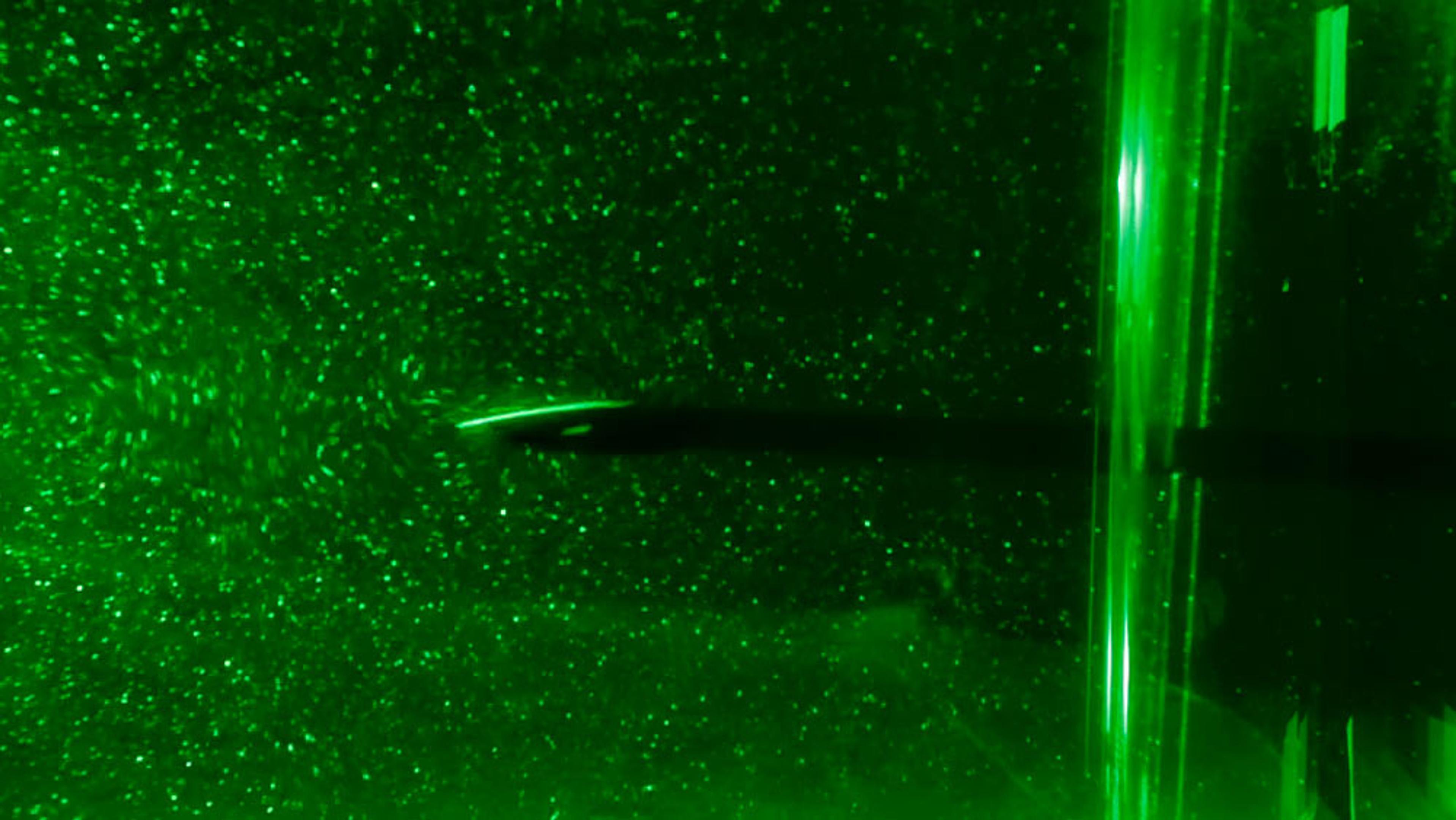 Photo showing particles in green liquid illuminated by a laser with a black object in the centre, demonstrating fluid dynamics.