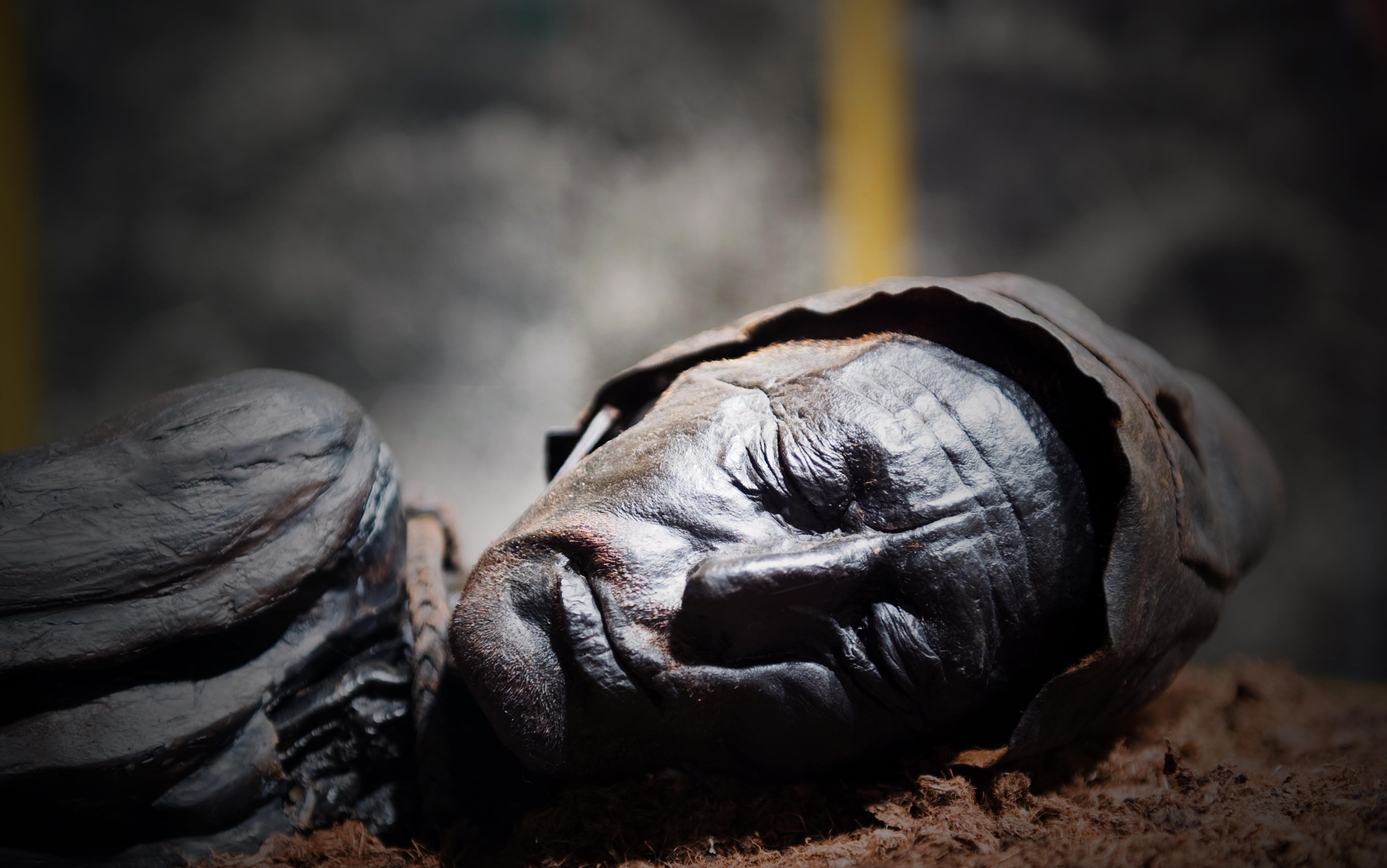 A well-preserved mummy with closed eyes, lying on a surface, wearing a cap. The background is blurred.