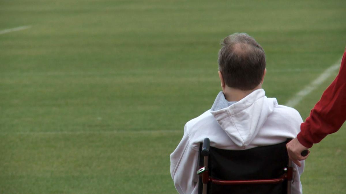 Photo of a man in a wheelchair on a grass field with another man’s hand resting on the wheelchair’s handle.