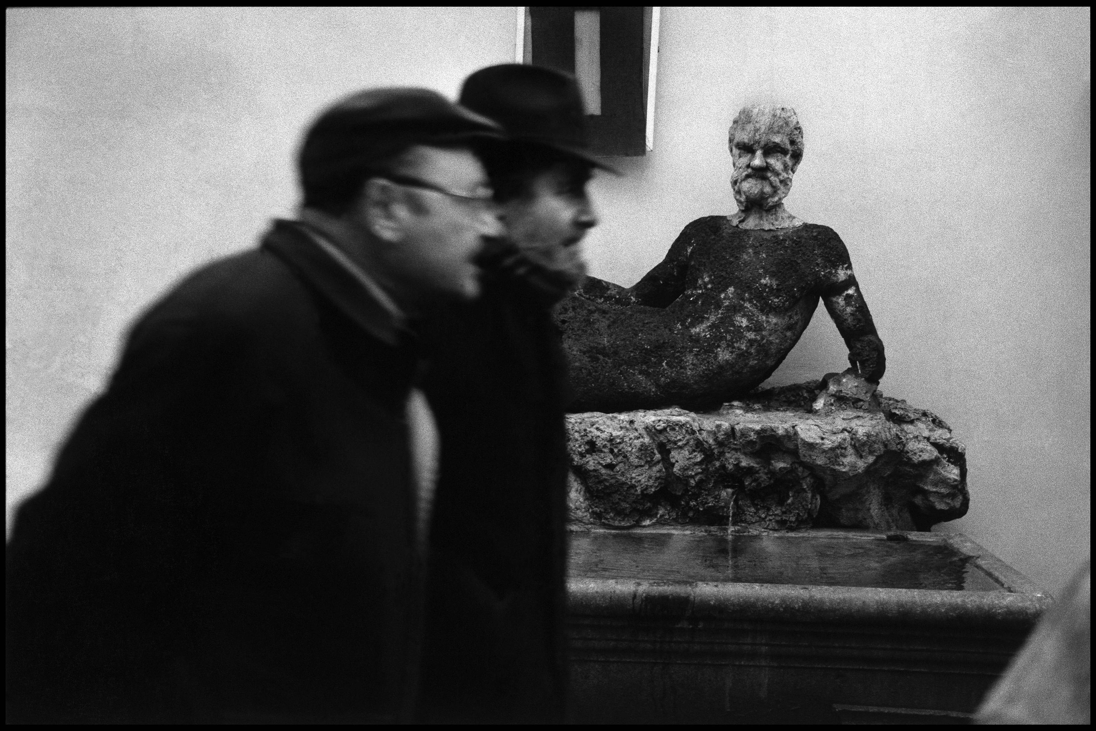 Black and white photo of two blurred men passing a weathered statue of a reclining man in a courtyard setting.