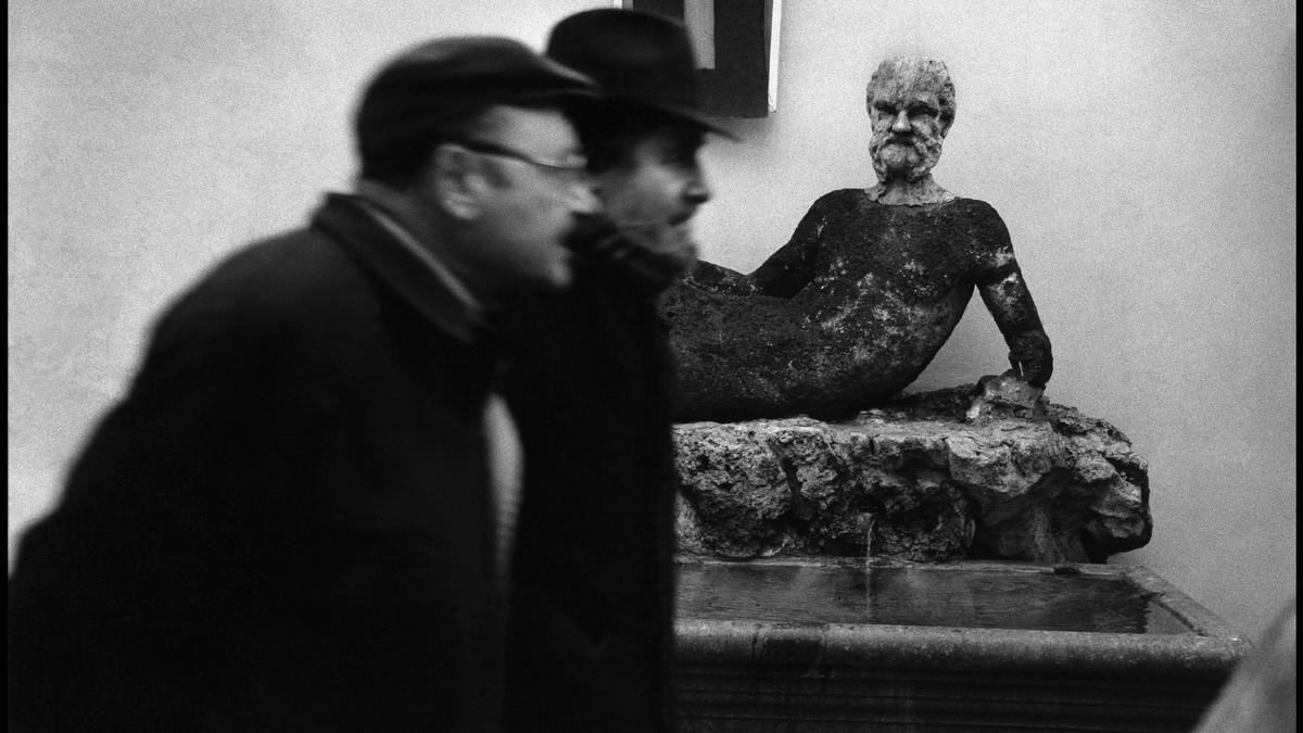 Black and white photo of two blurred men passing a weathered statue of a reclining man in a courtyard setting.
