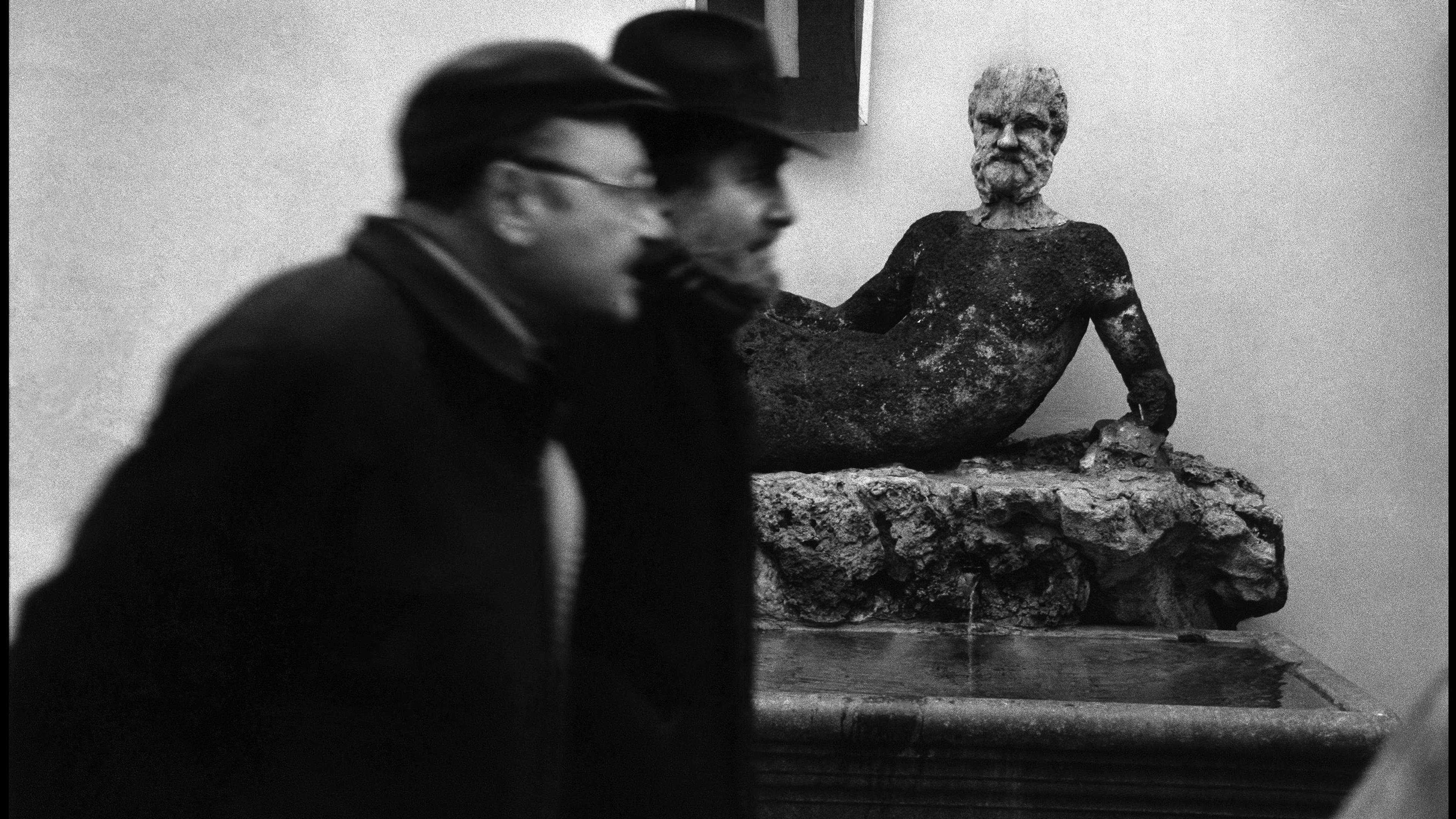 Black and white photo of two blurred men passing a weathered statue of a reclining man in a courtyard setting.