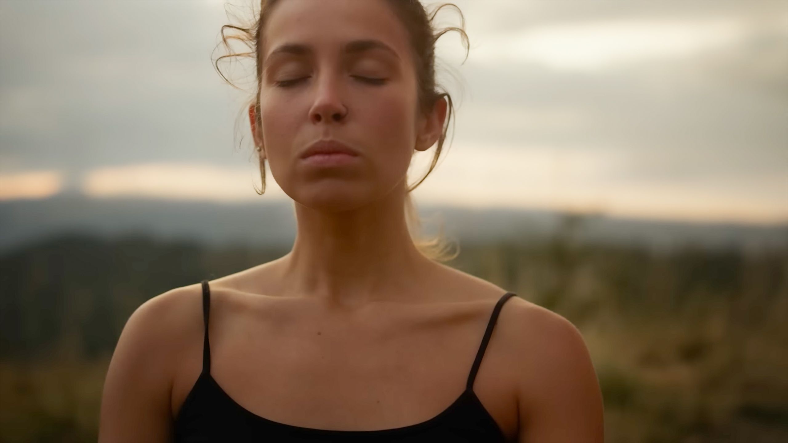 Photo of a serene woman outdoors with closed eyes in a black top, blurred landscape in the background.