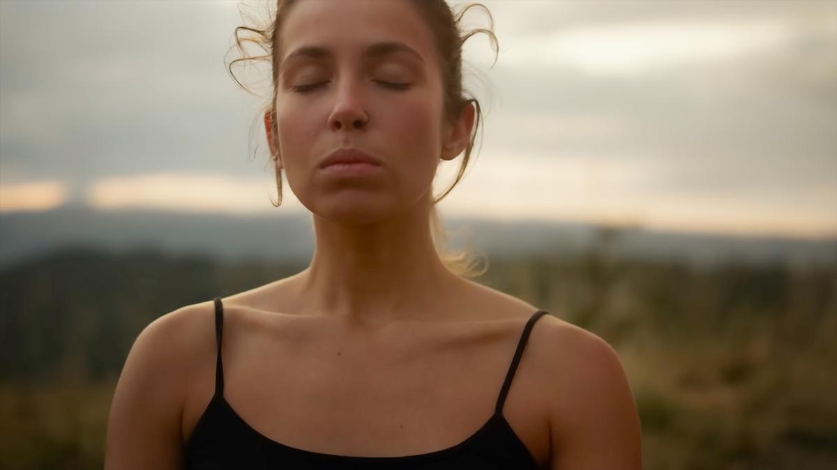 Photo of a serene woman outdoors with closed eyes in a black top, blurred landscape in the background.