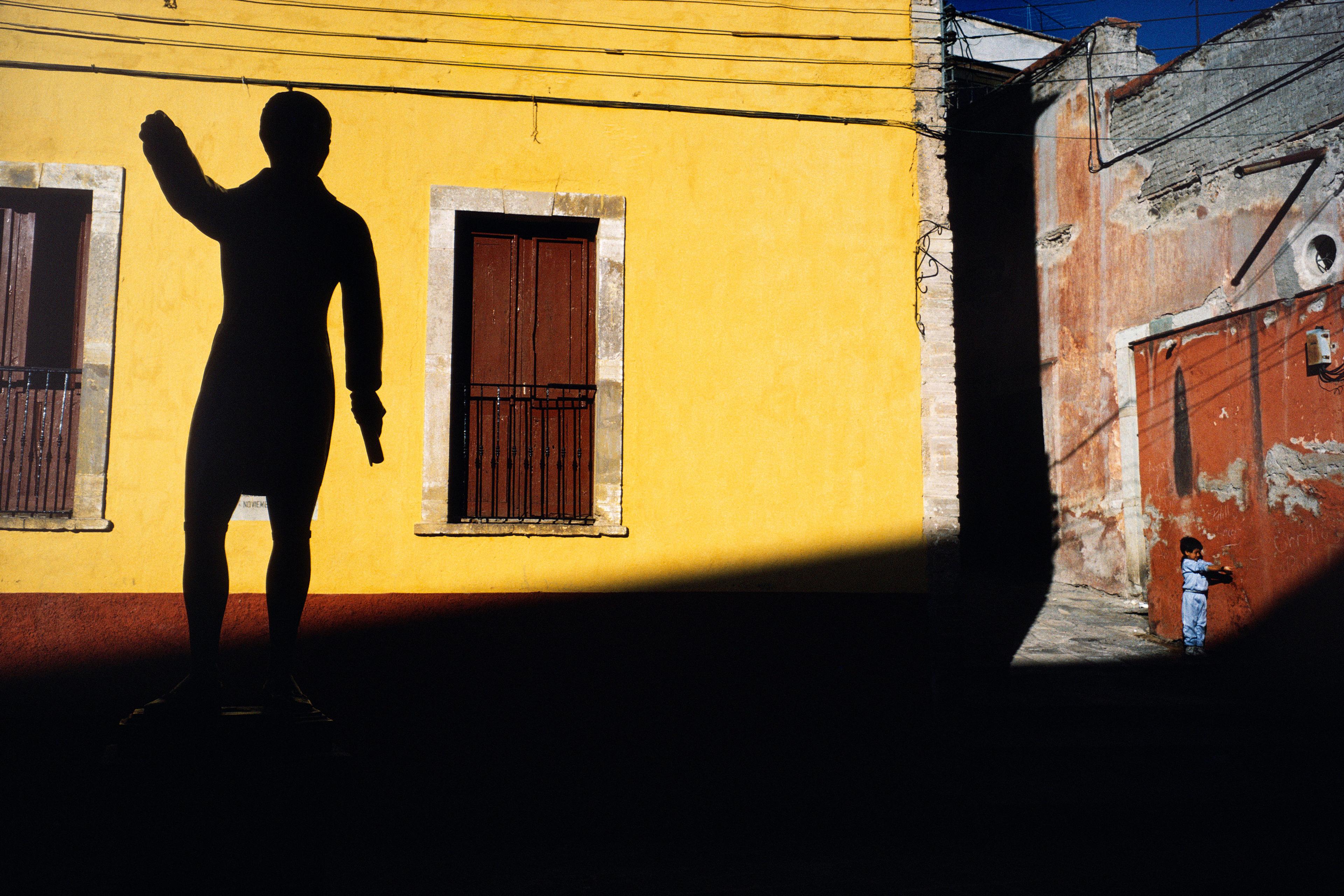 A large statue silhouetted on a yellow wall with a small boy in blue standing nearby on a sunlit street.
