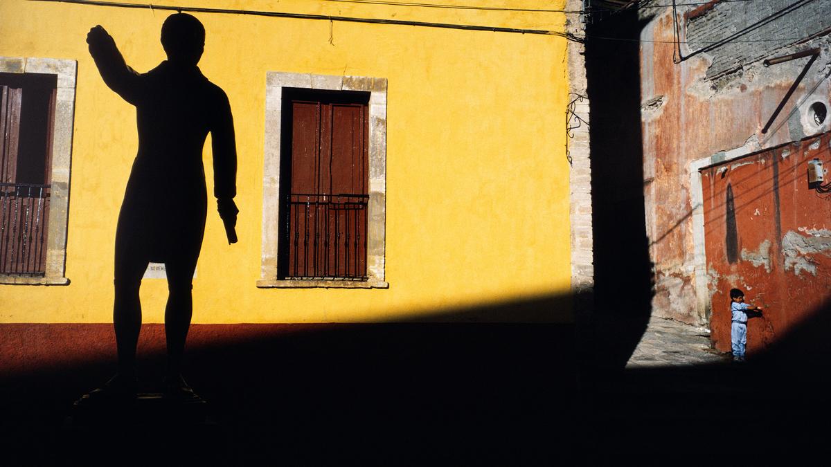 A large statue silhouetted on a yellow wall with a small boy in blue standing nearby on a sunlit street.