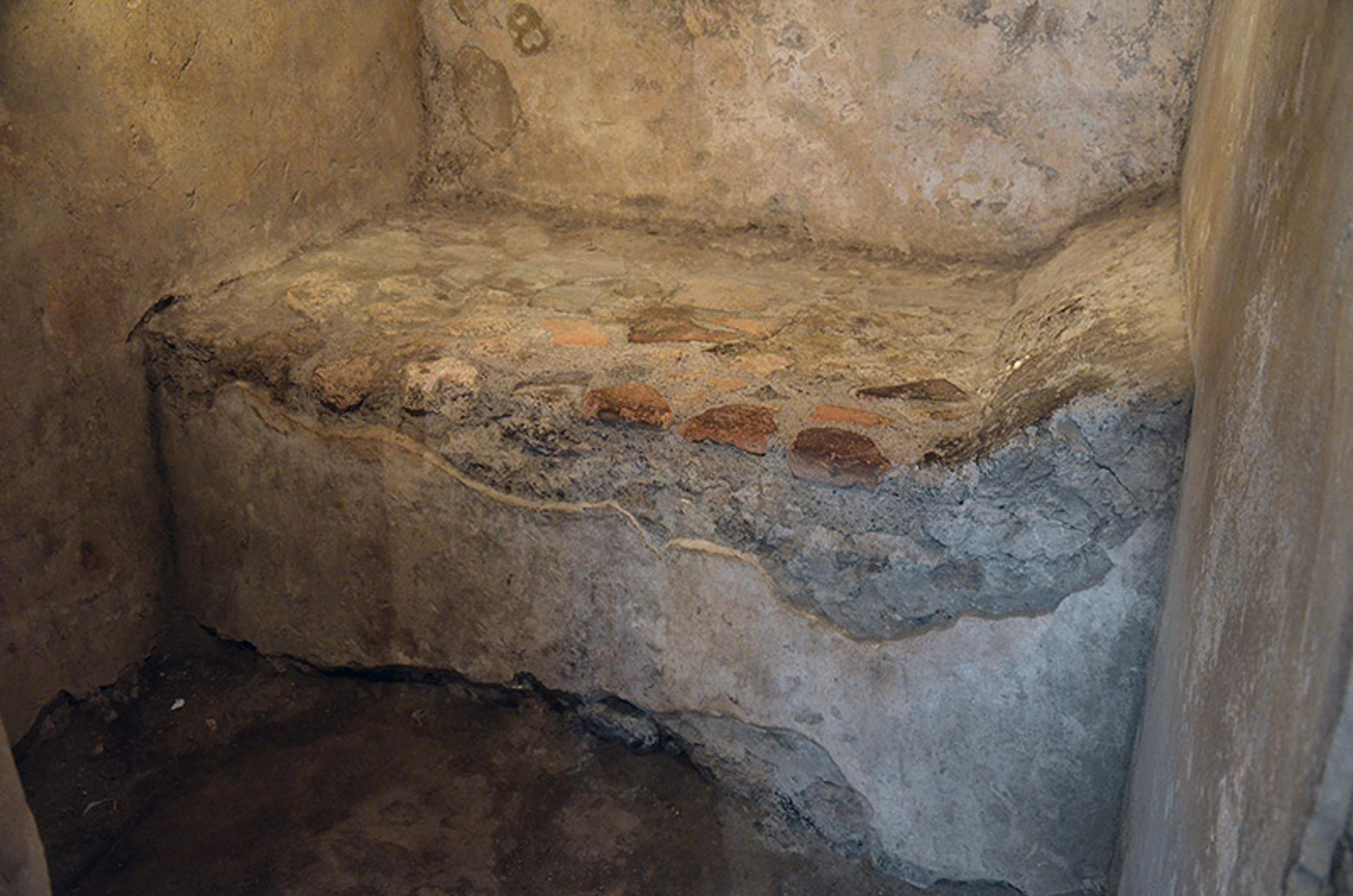 A rough, ancient stone seat built into a corner with weathered walls; the seat has patches of exposed red bricks.