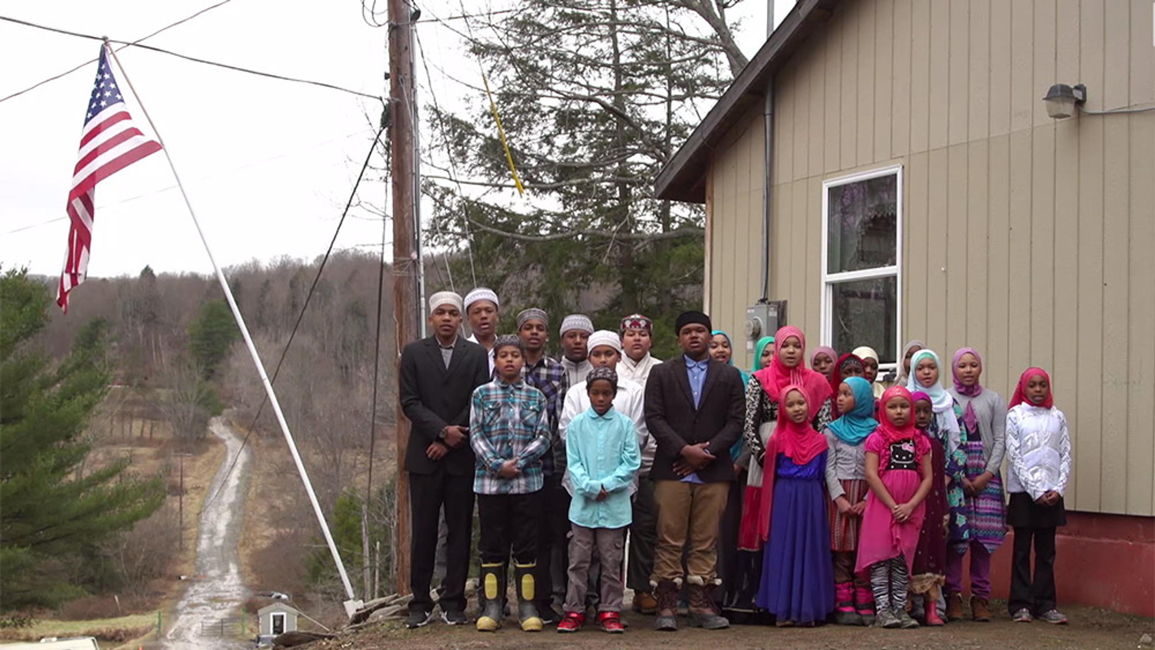 Members of an African-American Muslim community in Brooklyn, standing outside a building near a US flag with trees in the background.