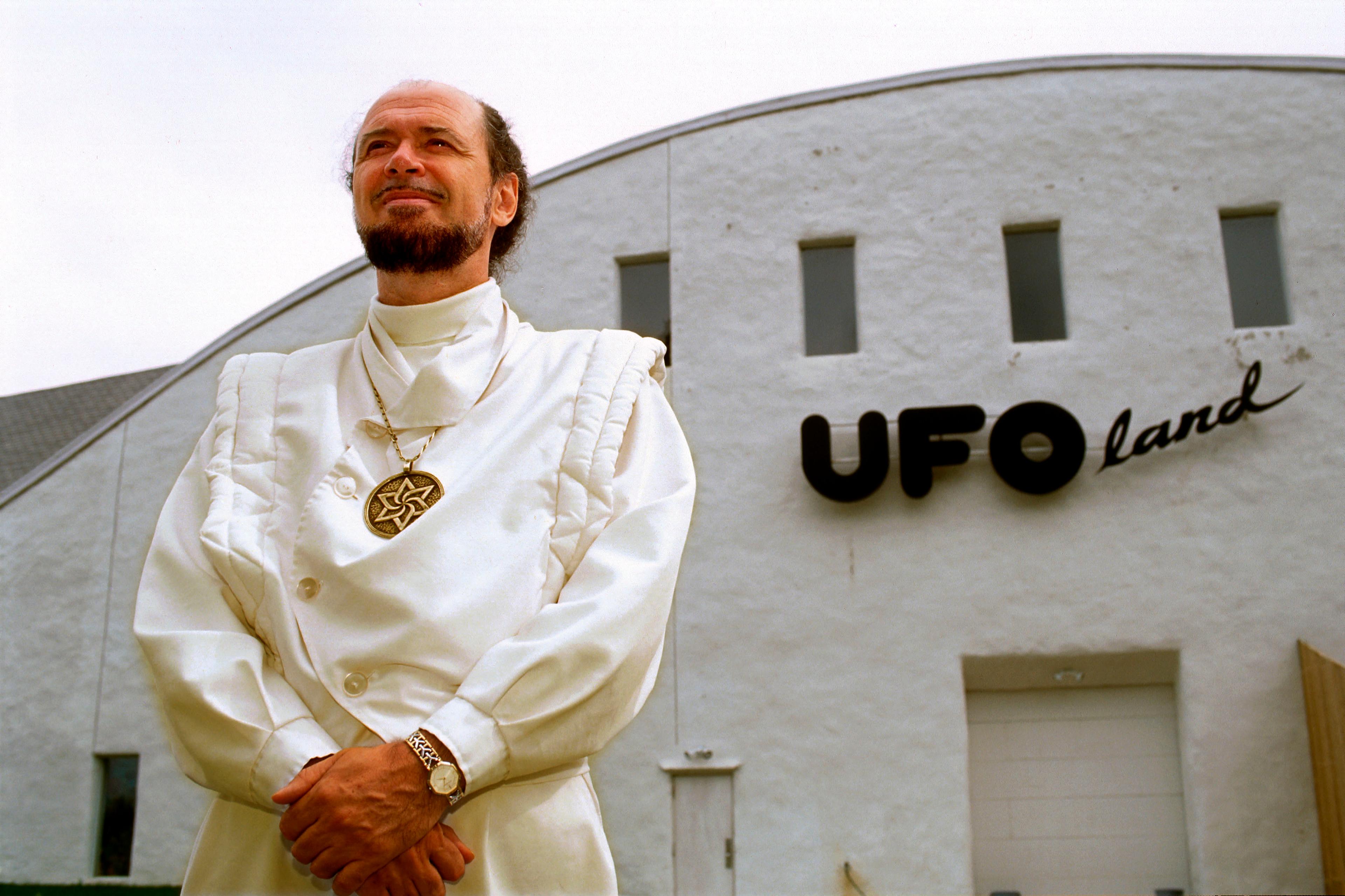 Photo of a man in white with a medallion standing in front of a building labelled “UFOland,” looking upwards confidently.
