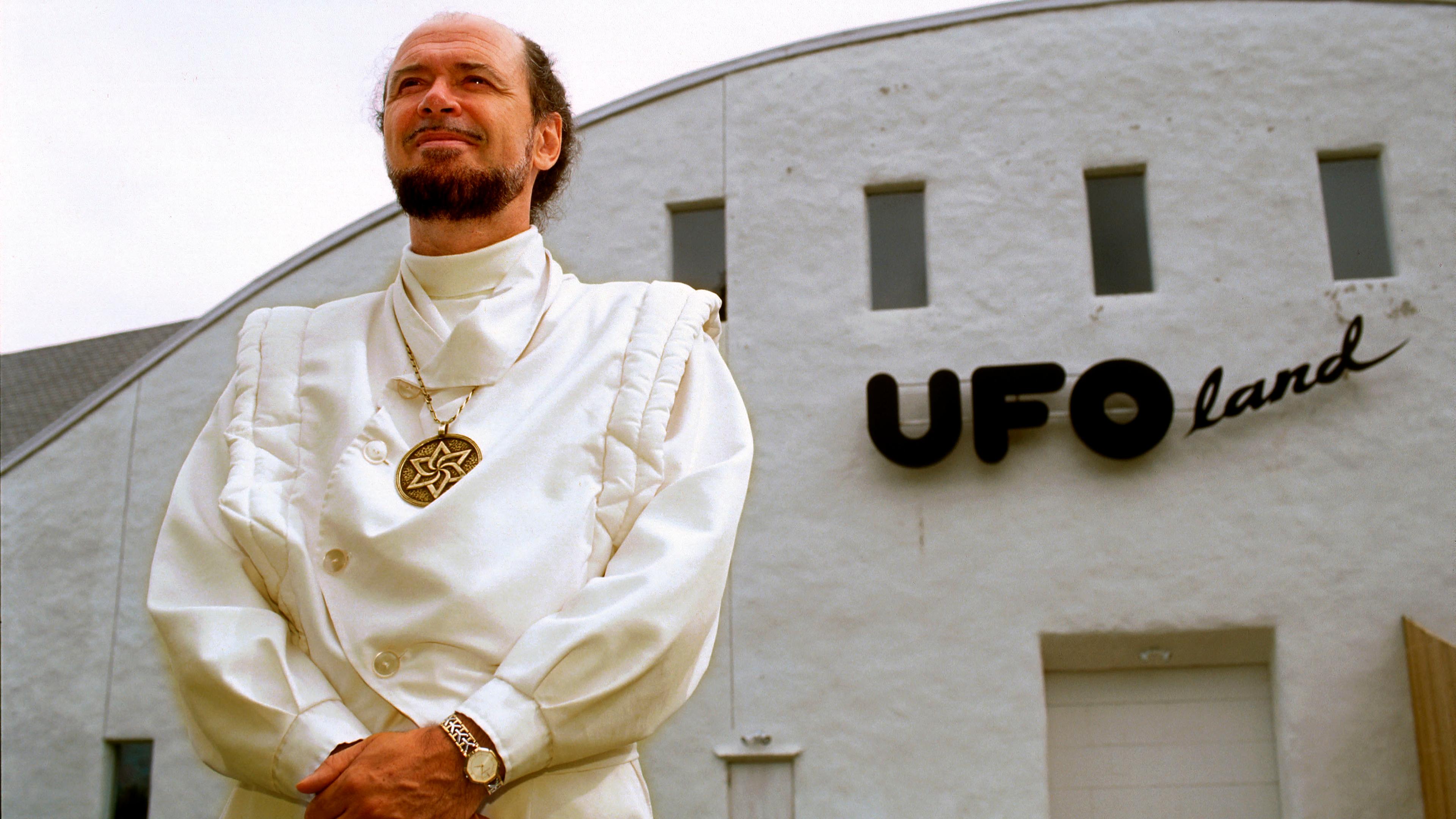 Photo of a man in white with a medallion standing in front of a building labelled “UFOland,” looking upwards confidently.