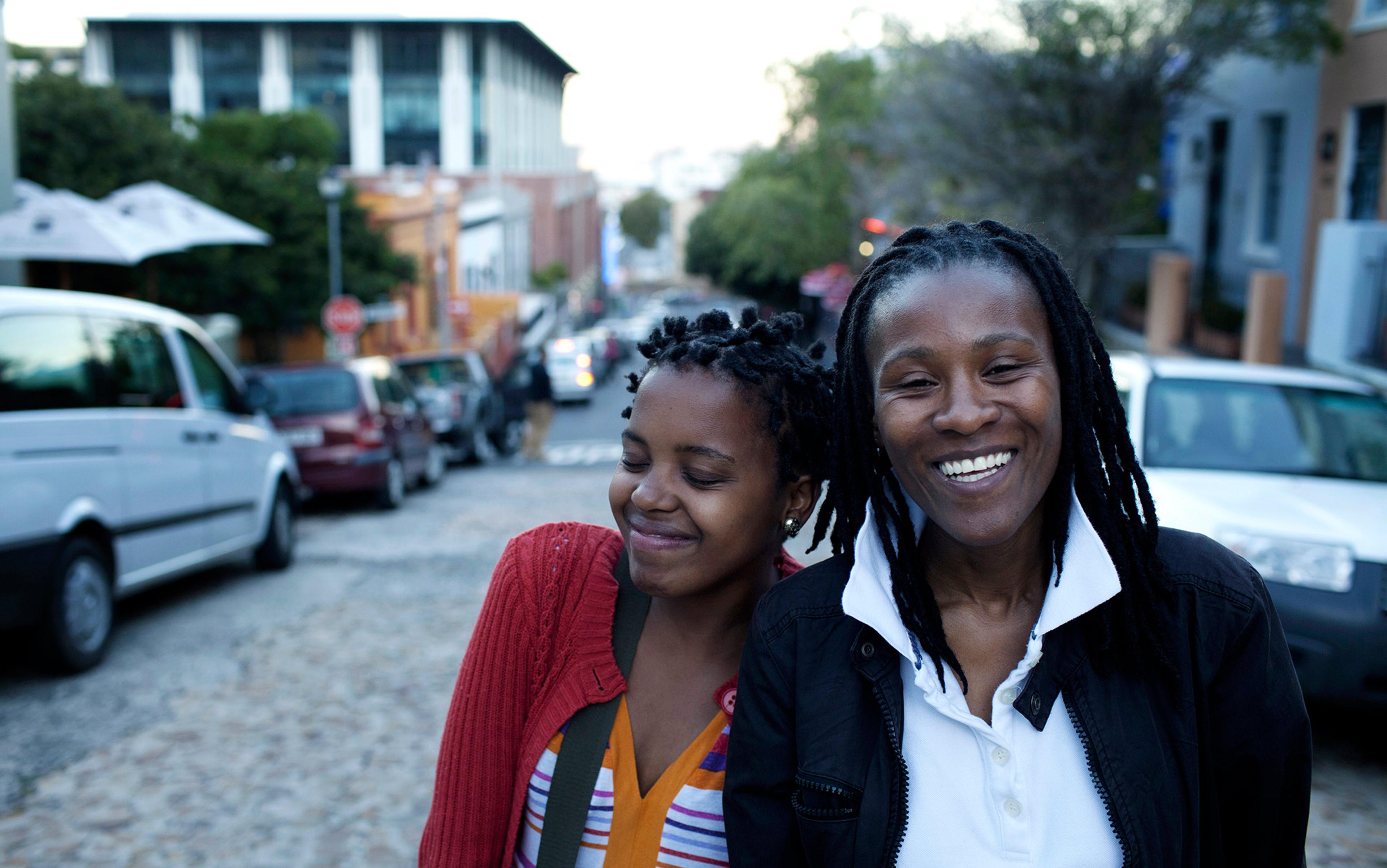 Two people smiling on a cobblestone street with parked cars and buildings in the background, taken during the day.