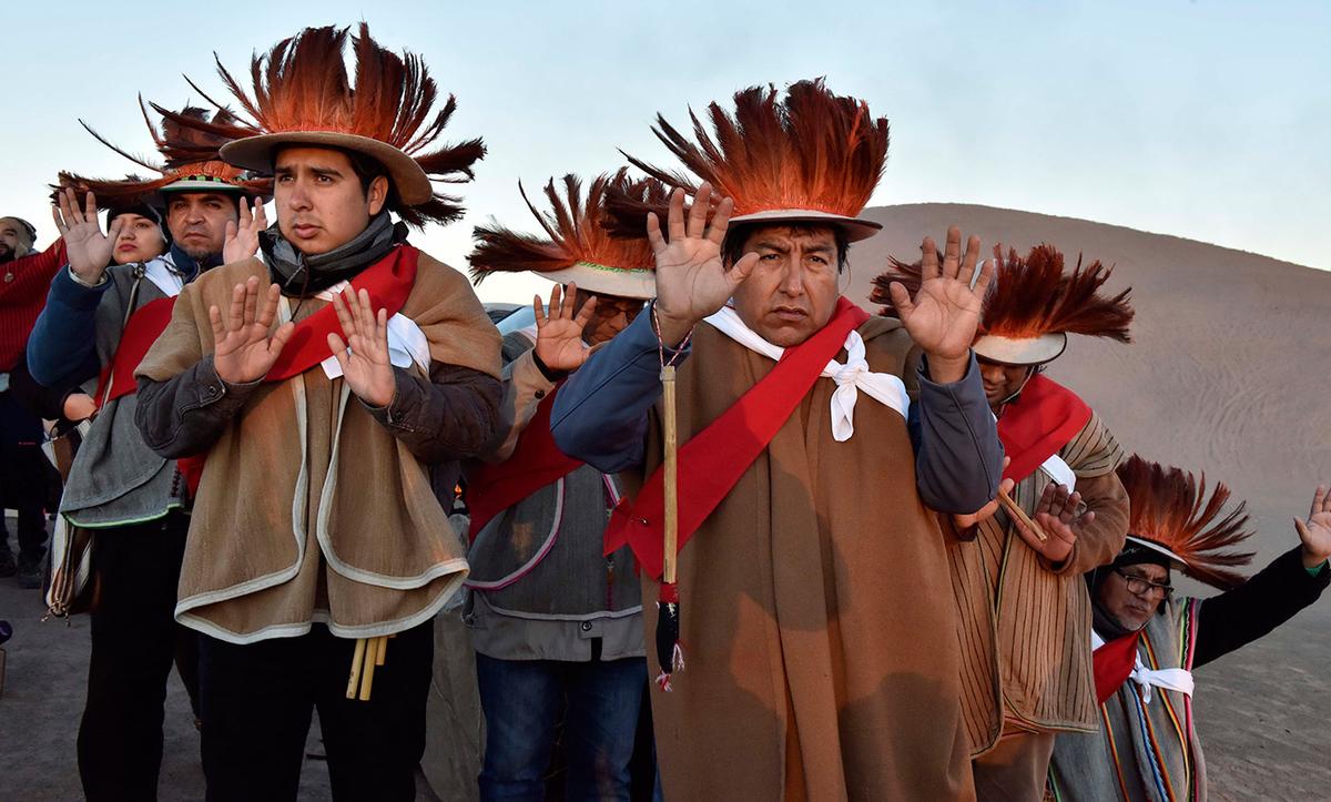 A group of people wearing traditional hats made of feathers, raising their hands in a ritual in a desert landscape.