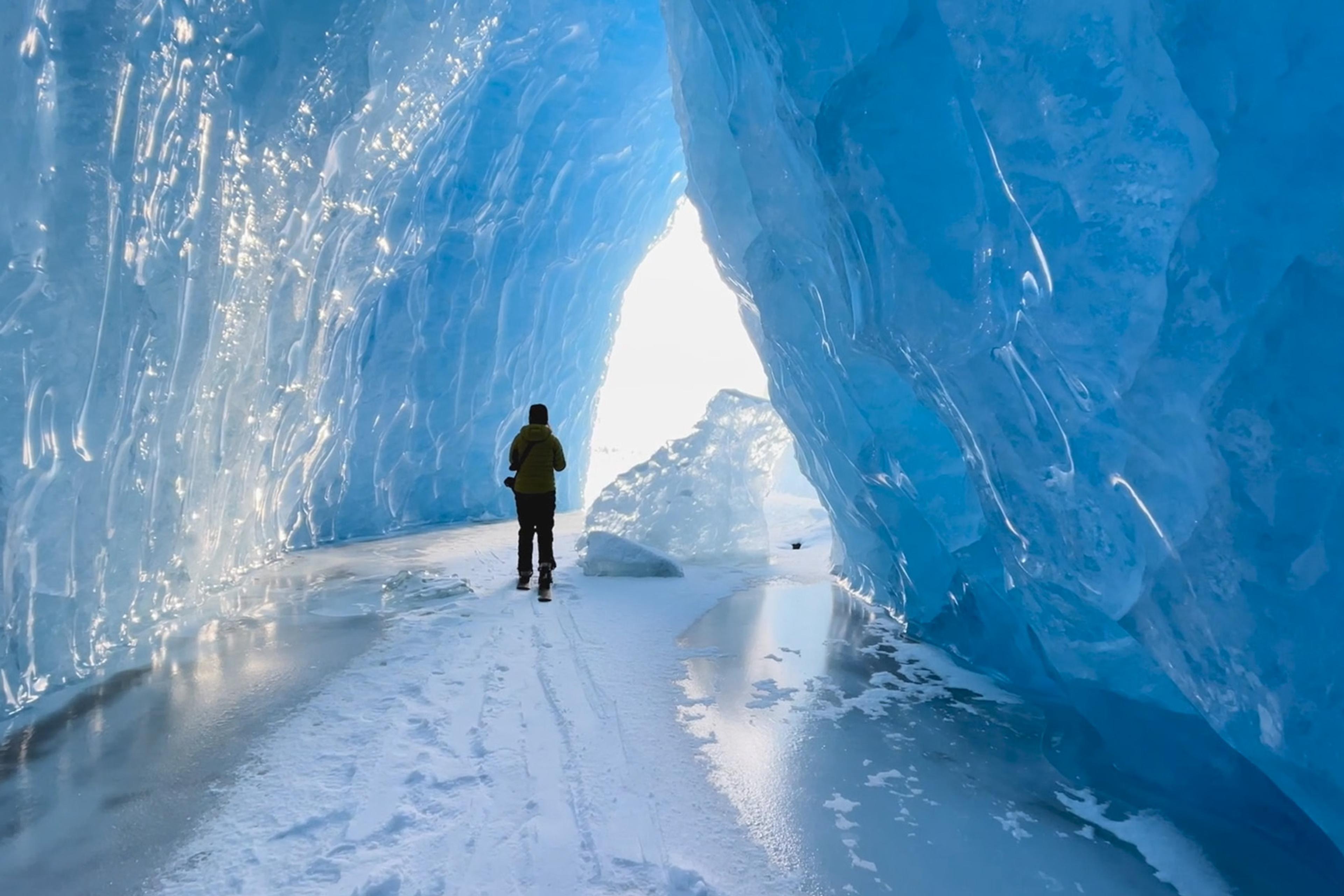 Person walking through a stunning ice cave with bright blue walls and a snow-covered ground, heading towards the light at the end.