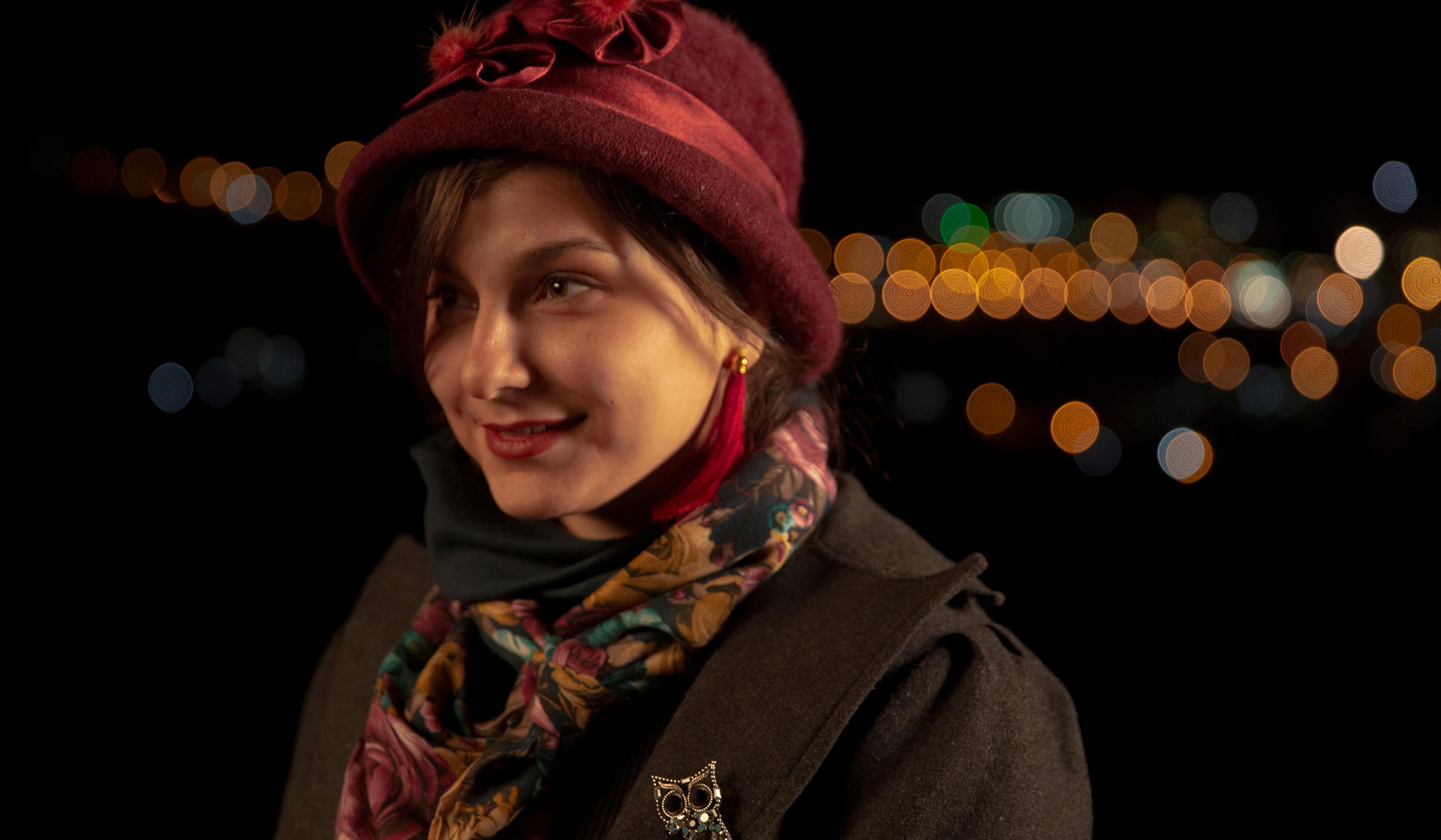 A woman wearing a maroon hat, earrings, a floral scarf, and an owl brooch, with city lights blurred in the background at night.