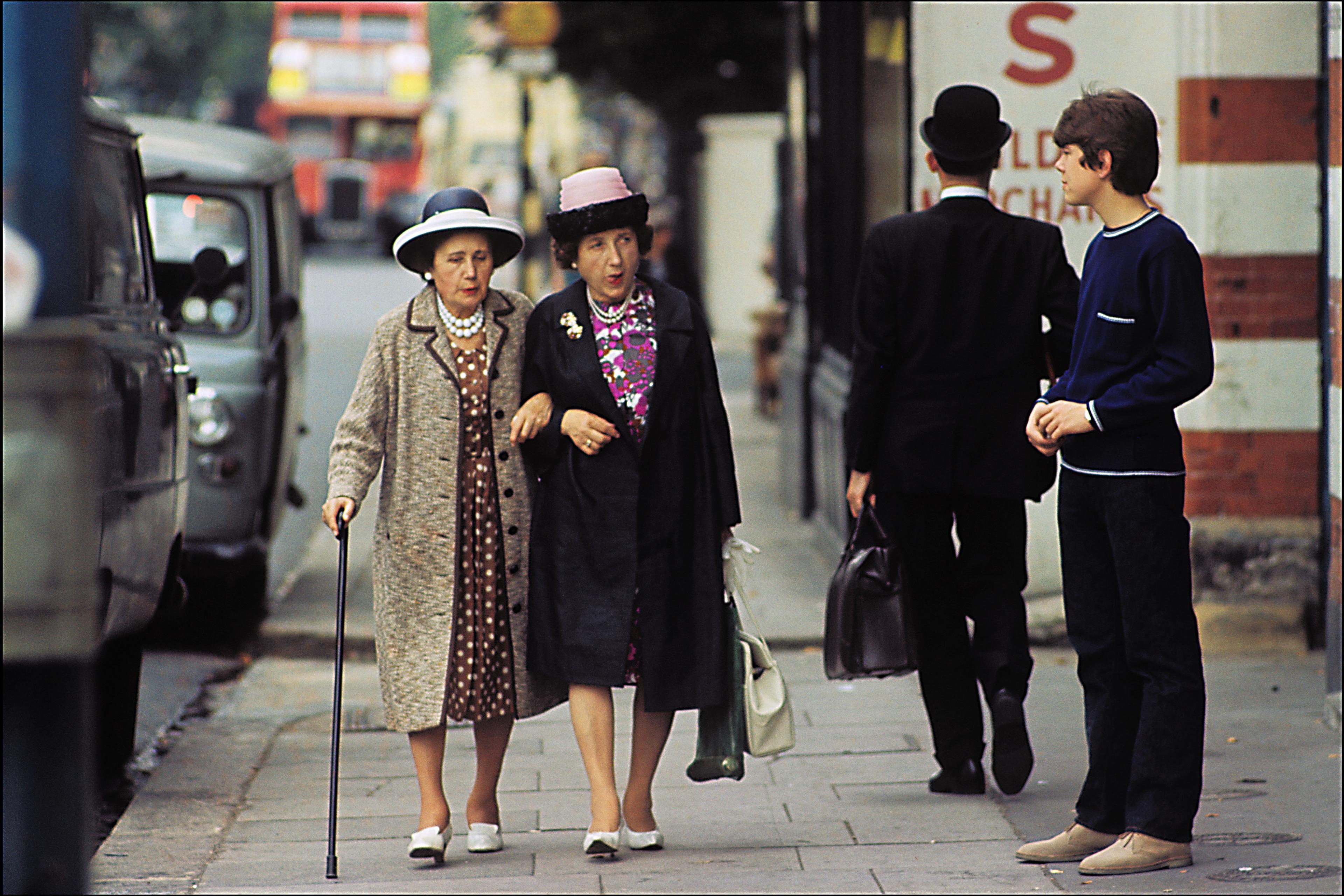 Photo of two elderly women walking together on a city pavement, with a red bus and two other people in the background.