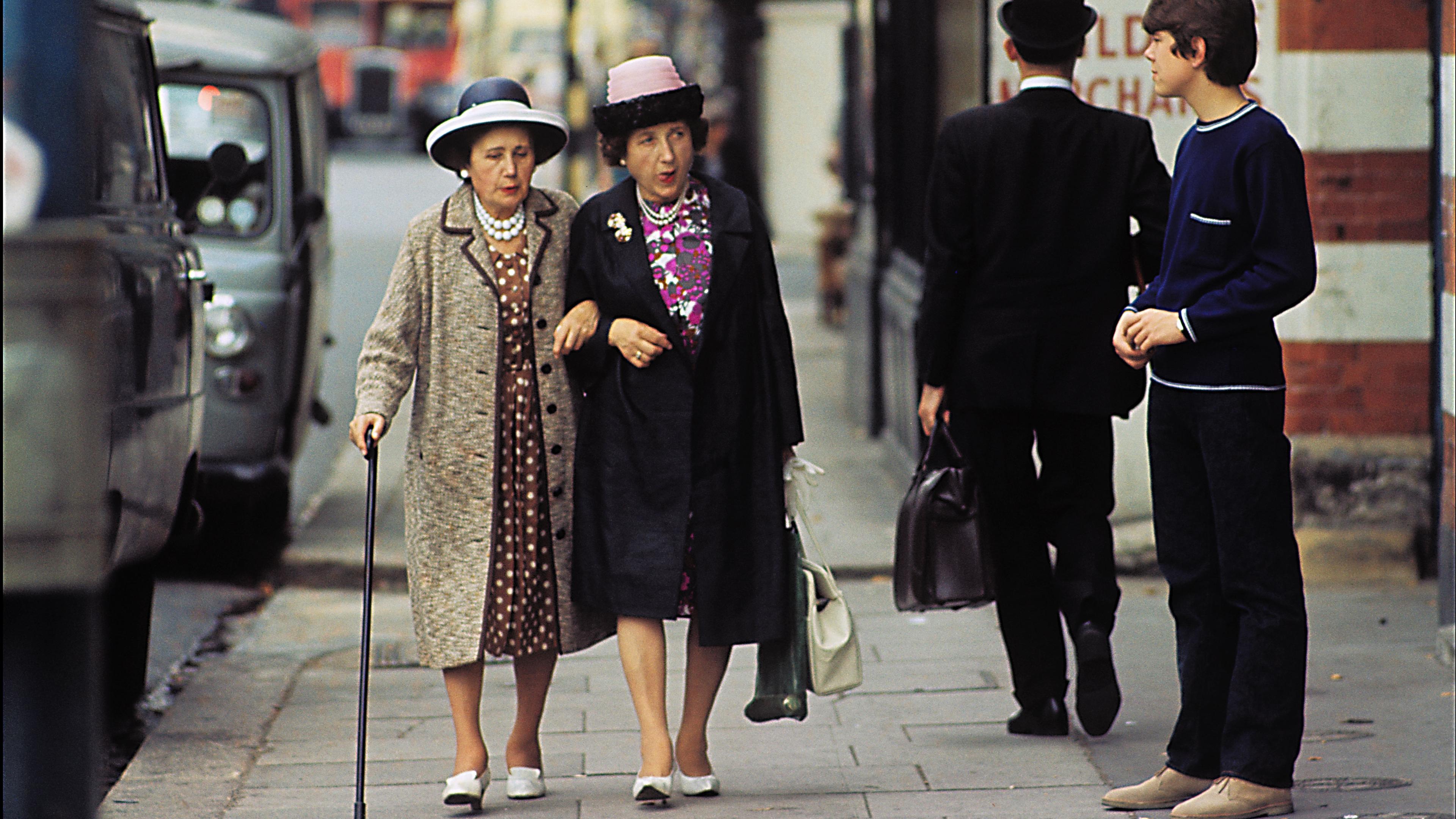 Photo of two elderly women walking together on a city pavement, with a red bus and two other people in the background.