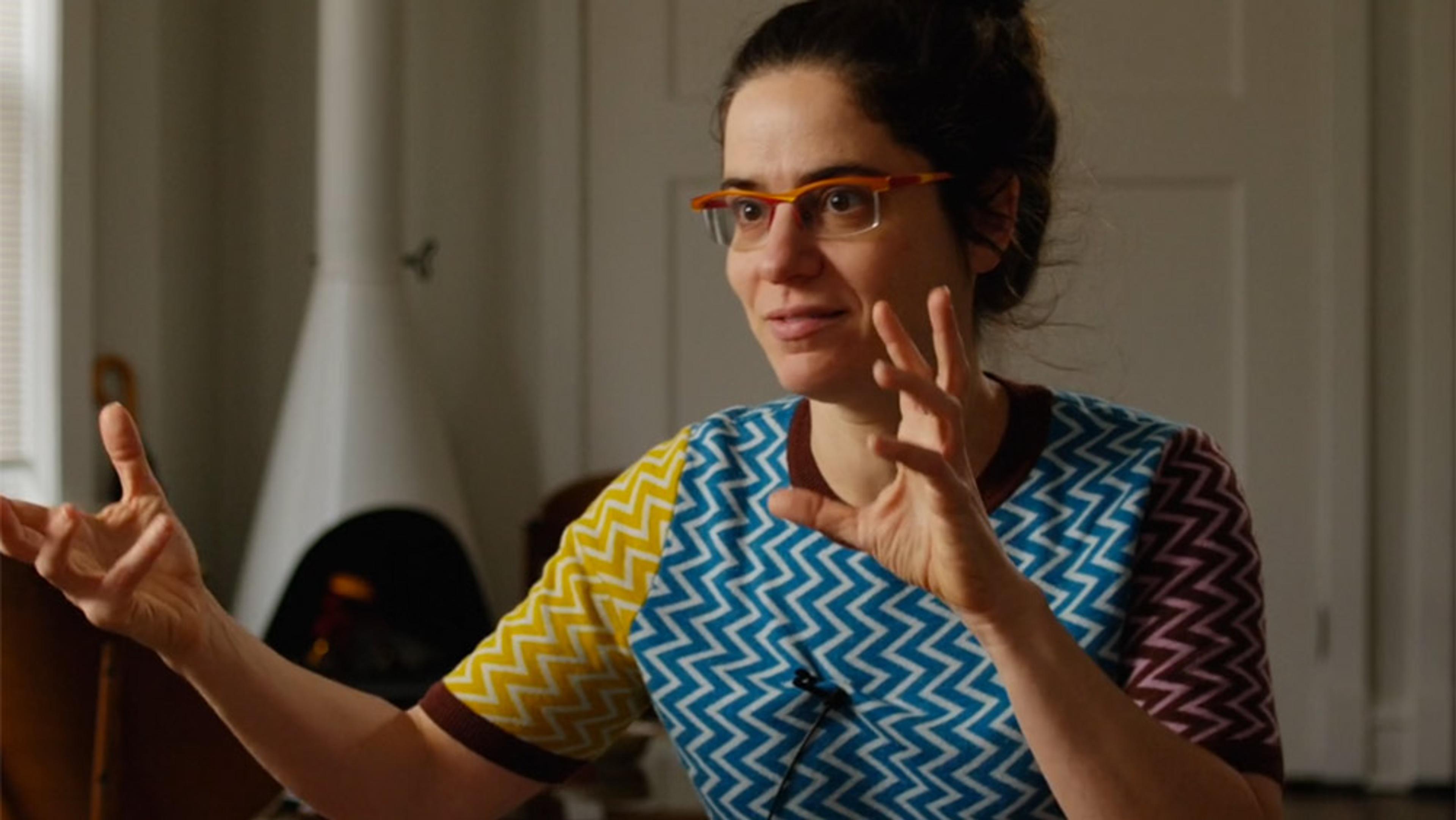 A woman with glasses wearing a colourful zigzag-patterned shirt who is Agnes Callard inside a room gesturing with her hands.