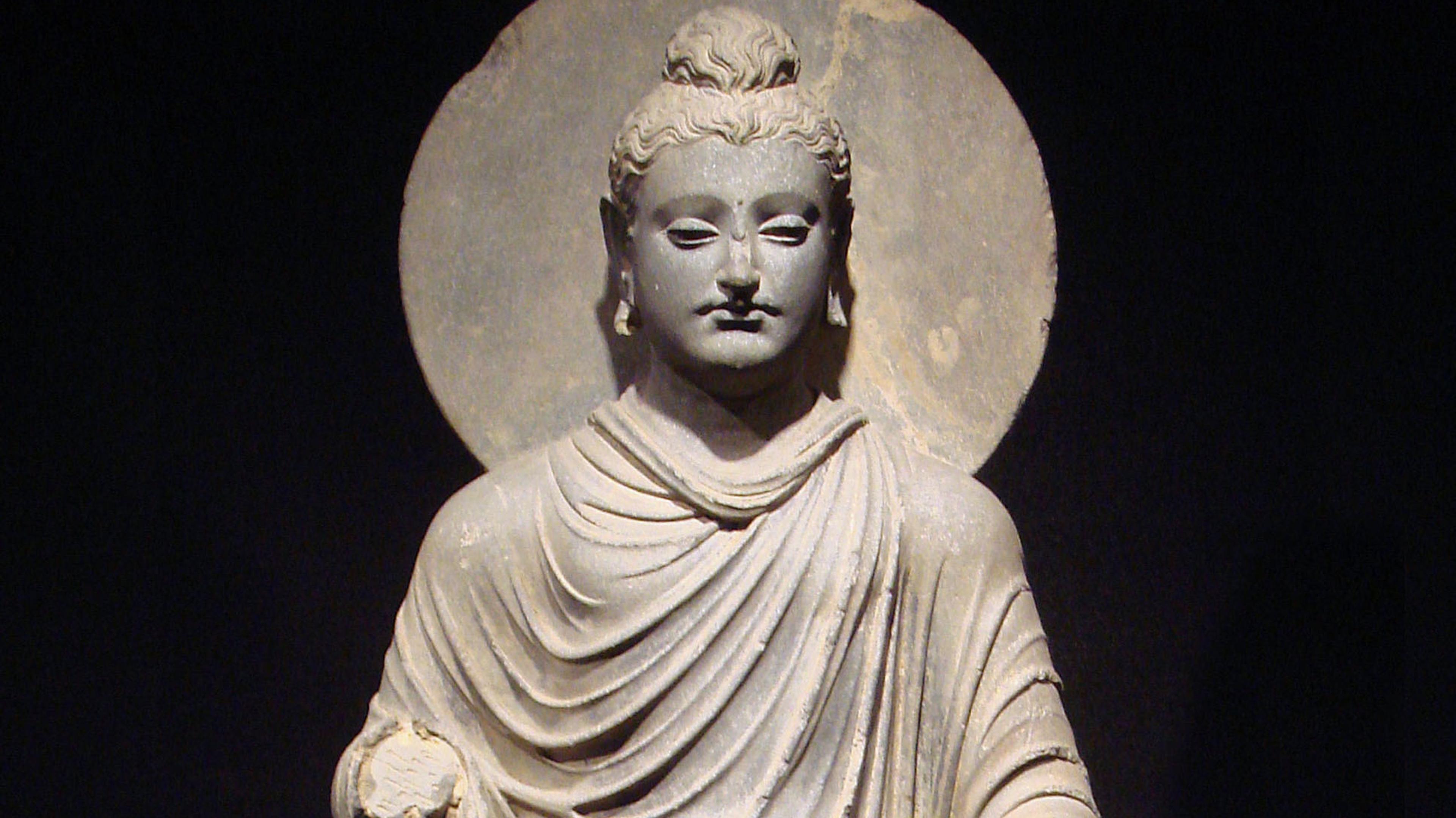 Stone sculpture of a serene figure in draped robes with a halo against a dark background.