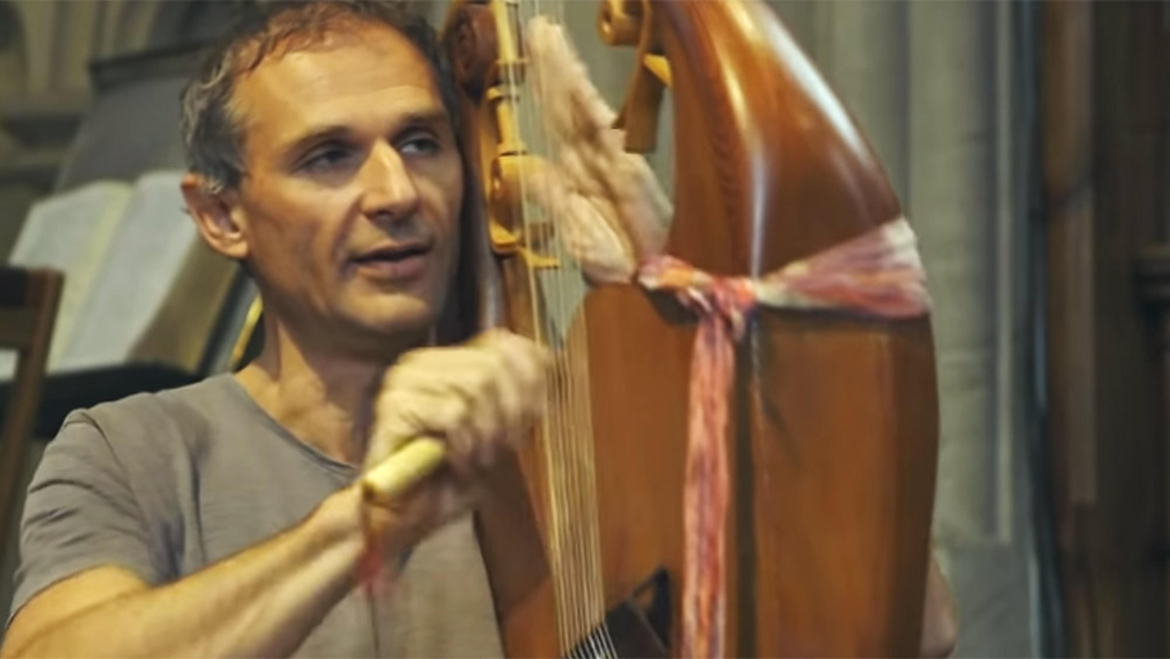 A man playing a string instrument with focus on his face and hands, with books and furniture in the background.
