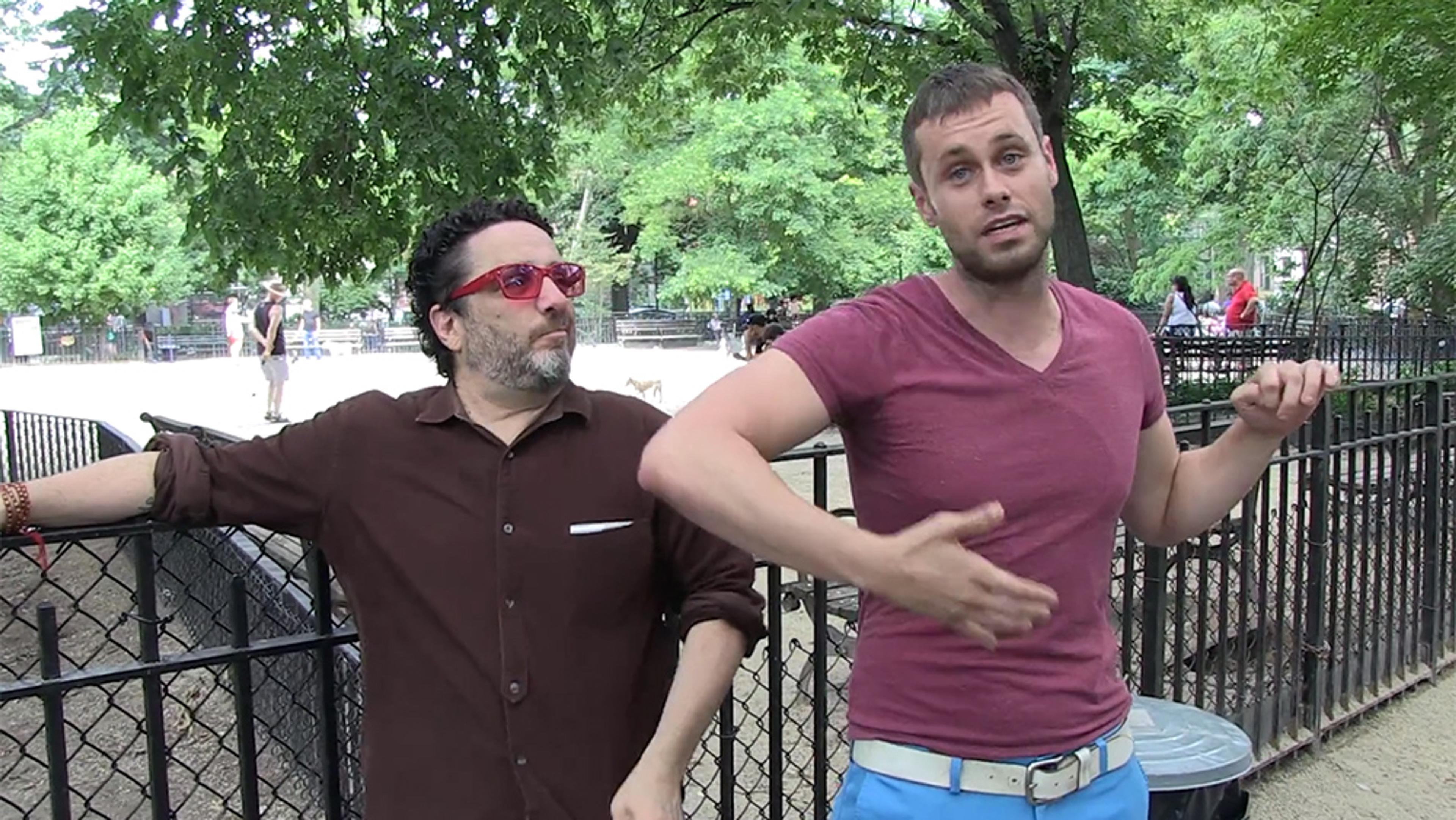 Two men standing by a fence in a park; one wears a brown shirt and red glasses, the other a maroon shirt and blue shorts.