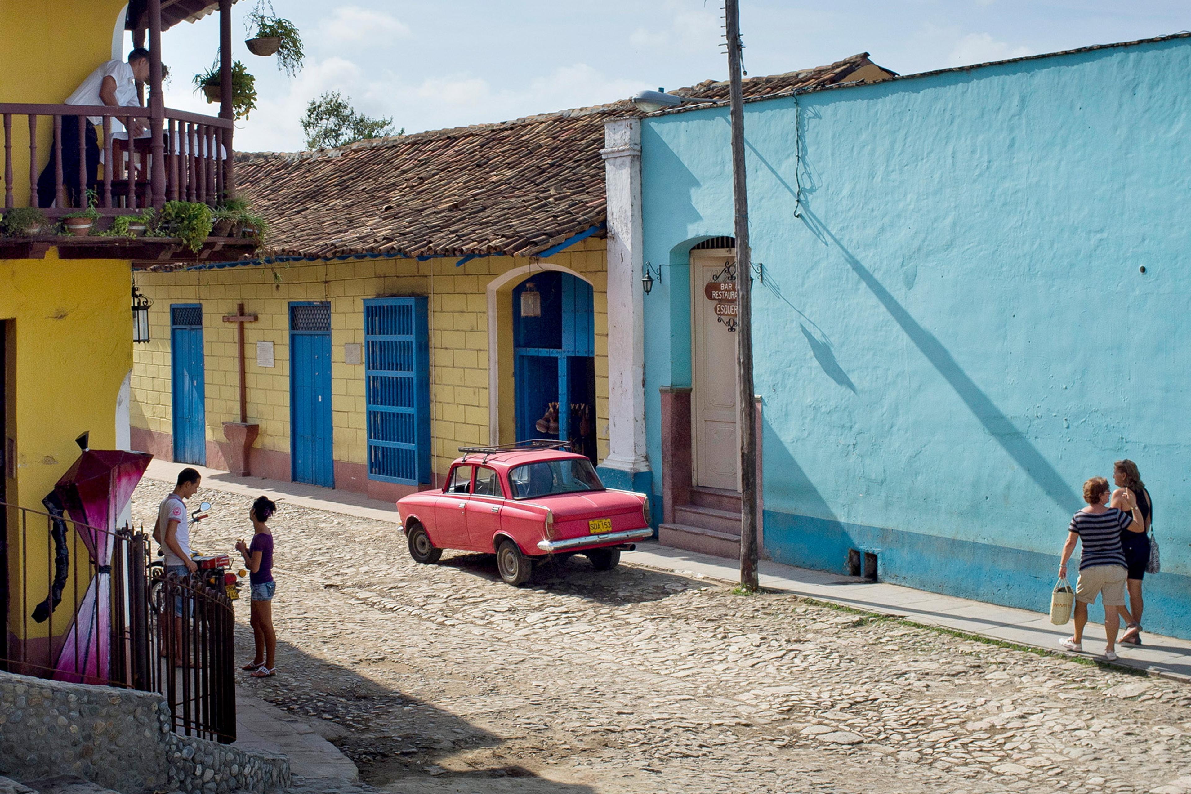 Photo of a cobblestone street with colourful buildings and a red car. People are walking and talking on a sunny day.