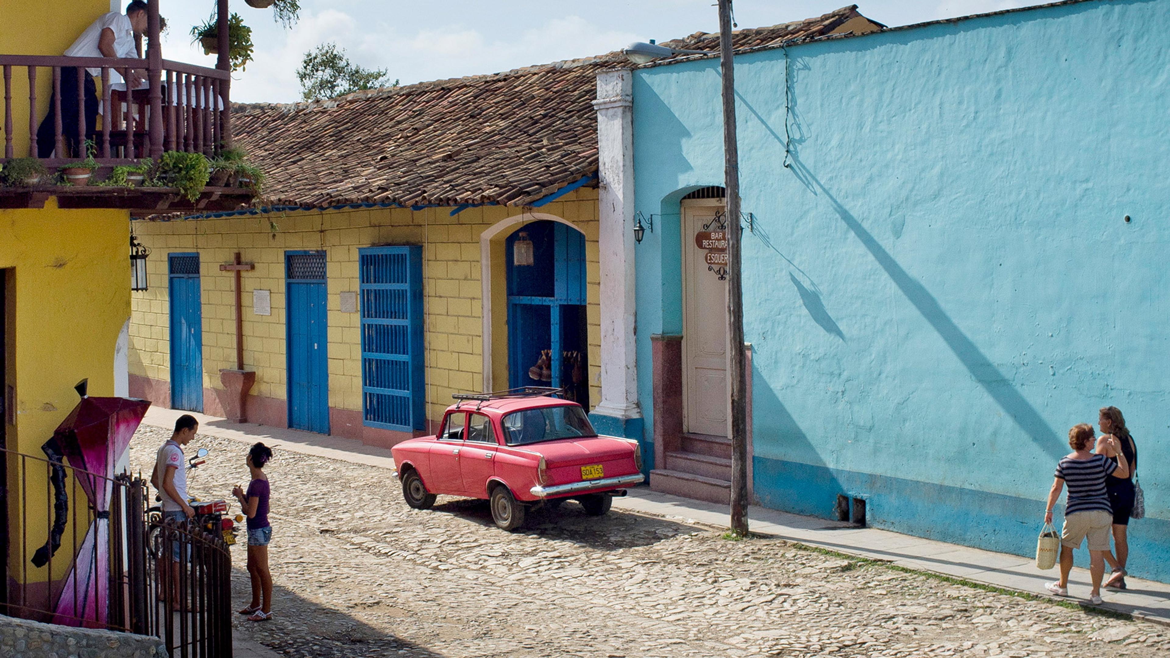 Photo of a cobblestone street with colourful buildings and a red car. People are walking and talking on a sunny day.