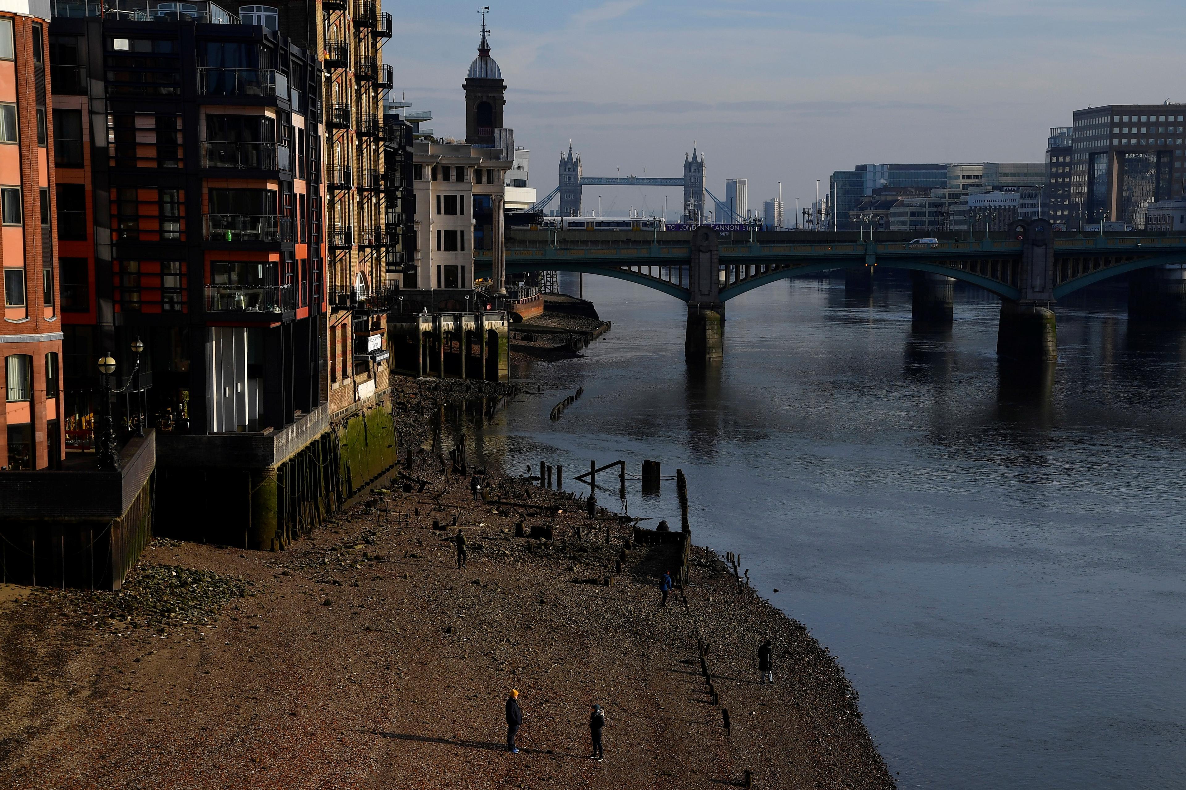 Photo of a riverside with buildings, a bridge and Tower Bridge in view, pedestrians on a pebbled shore in the foreground.