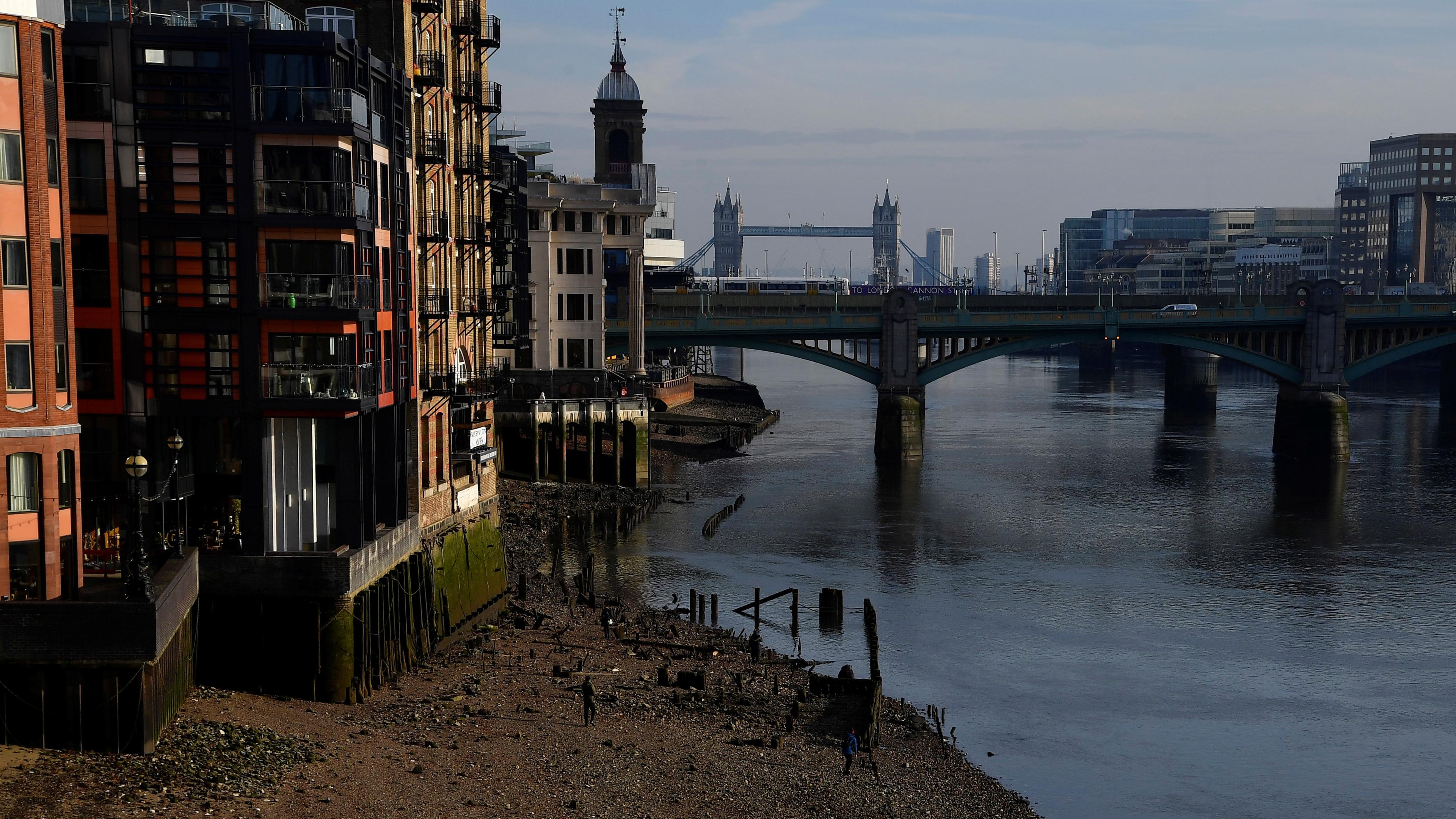 Photo of a riverside with buildings, a bridge and Tower Bridge in view, pedestrians on a pebbled shore in the foreground.