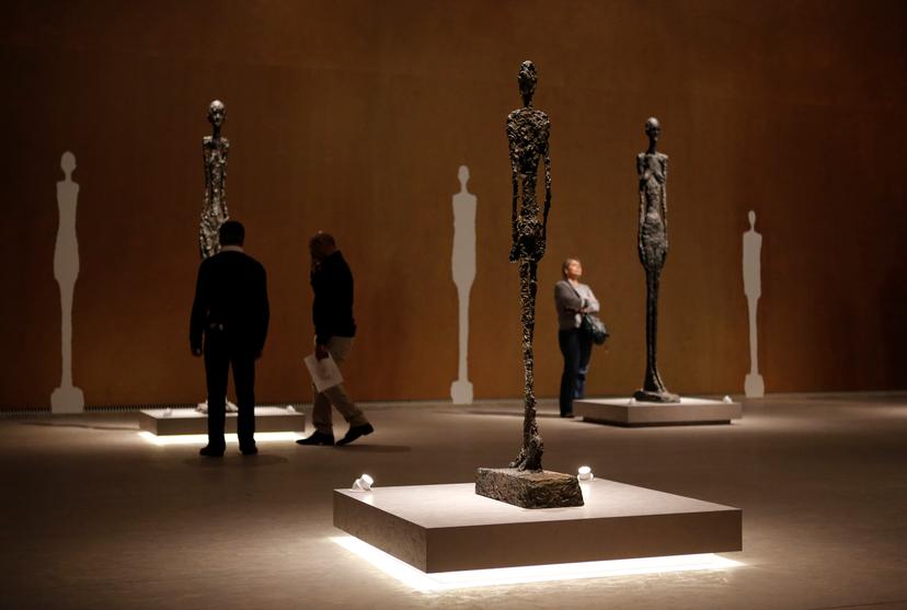 Photo of a museum exhibition with three tall, thin sculptures on pedestals and people observing them, with white shadows of the figures painted on the background wall.