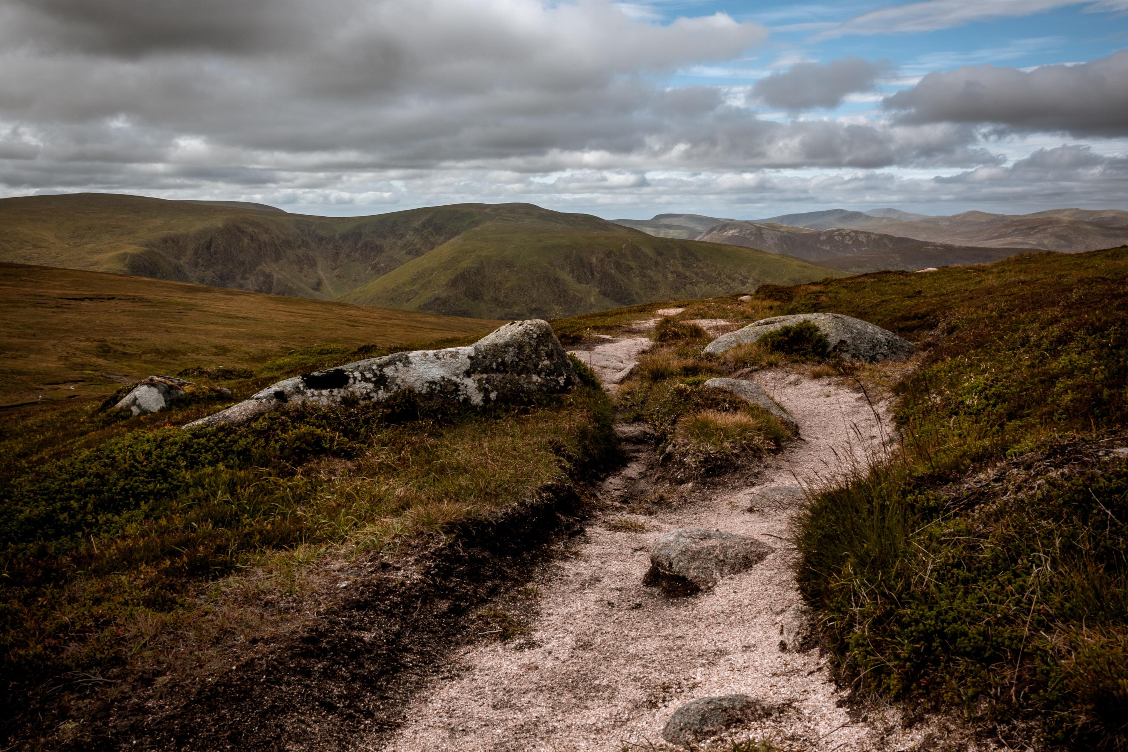 Photo of a rocky path winding through a grassy landscape with cloudy skies and distant hills in the background.