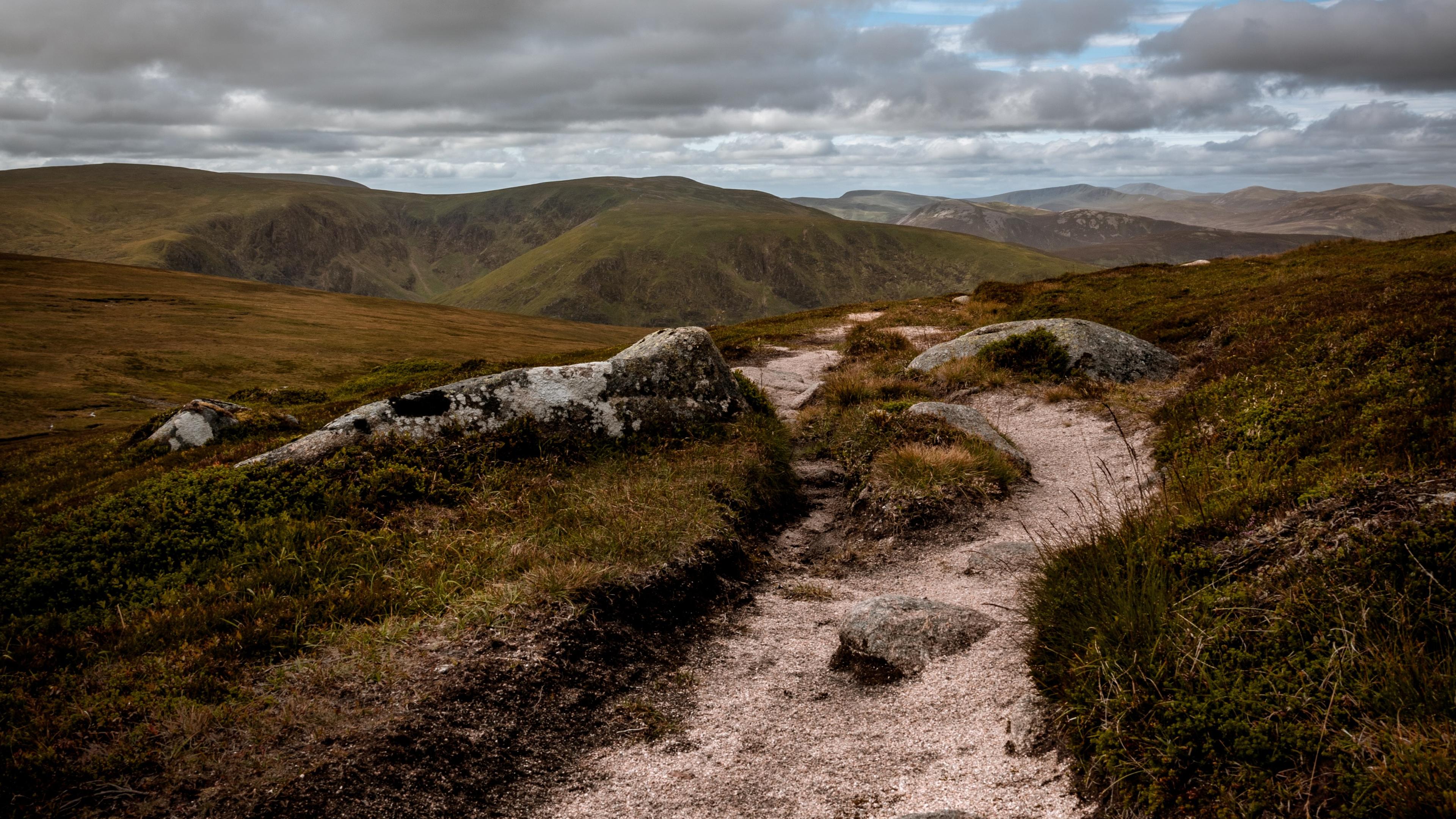 Photo of a rocky path winding through a grassy landscape with cloudy skies and distant hills in the background.