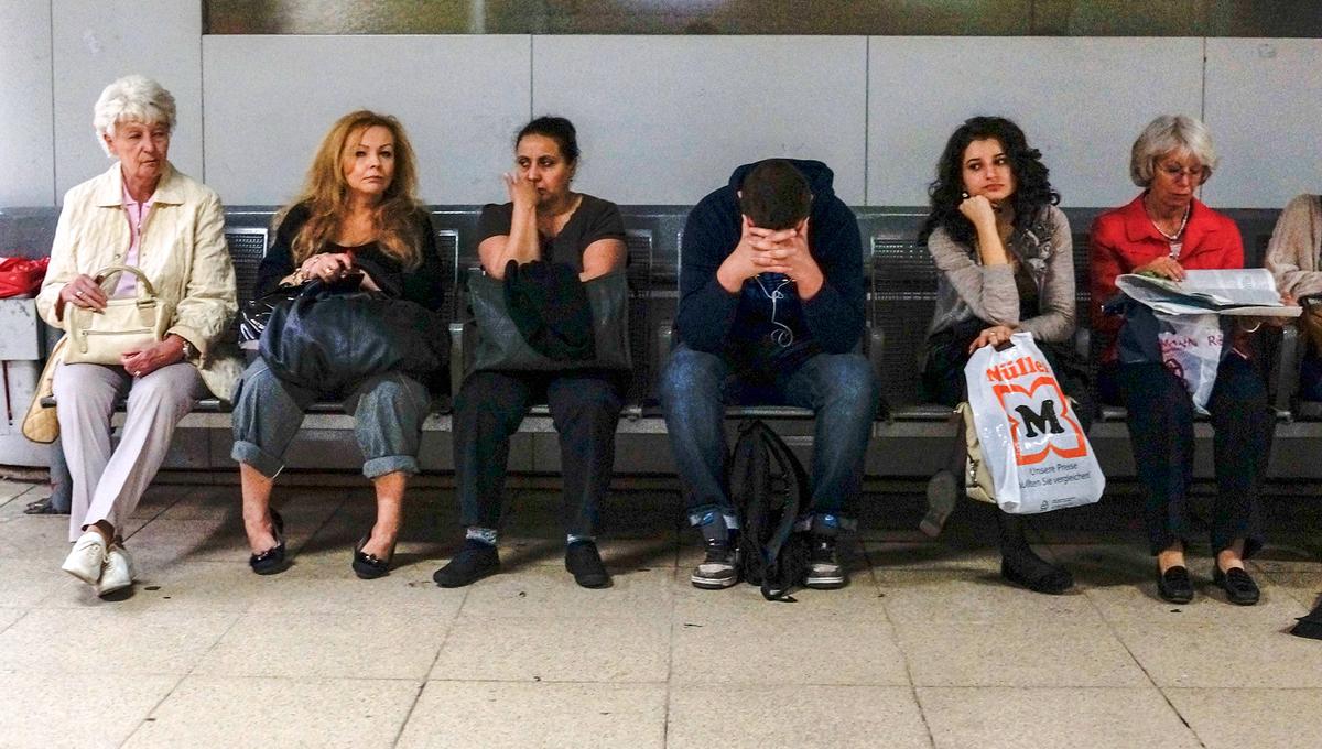 Photo of six people sitting on a bench in a waiting area appearing bored or pensive.