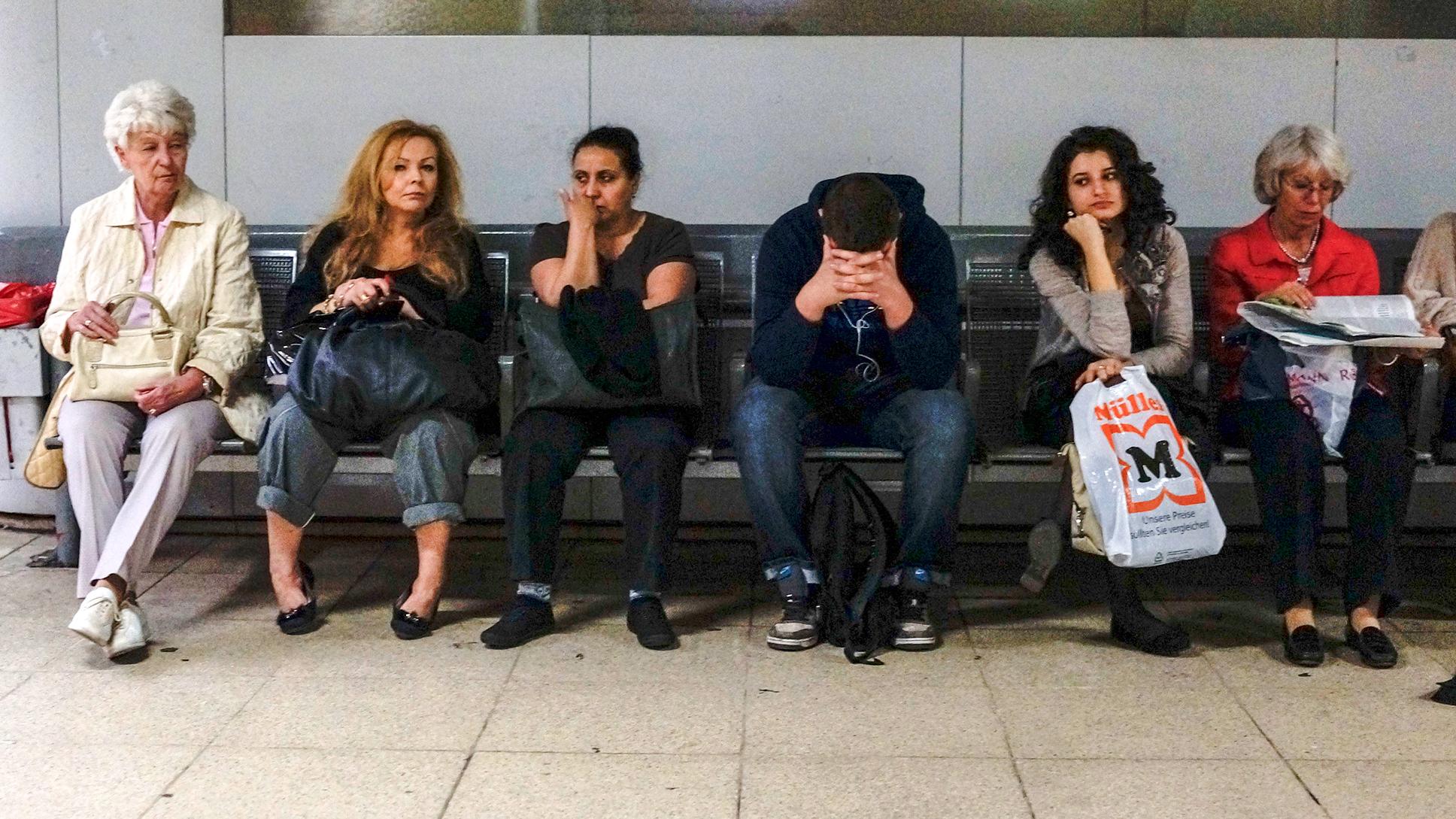 Photo of six people sitting on a bench in a waiting area appearing bored or pensive.