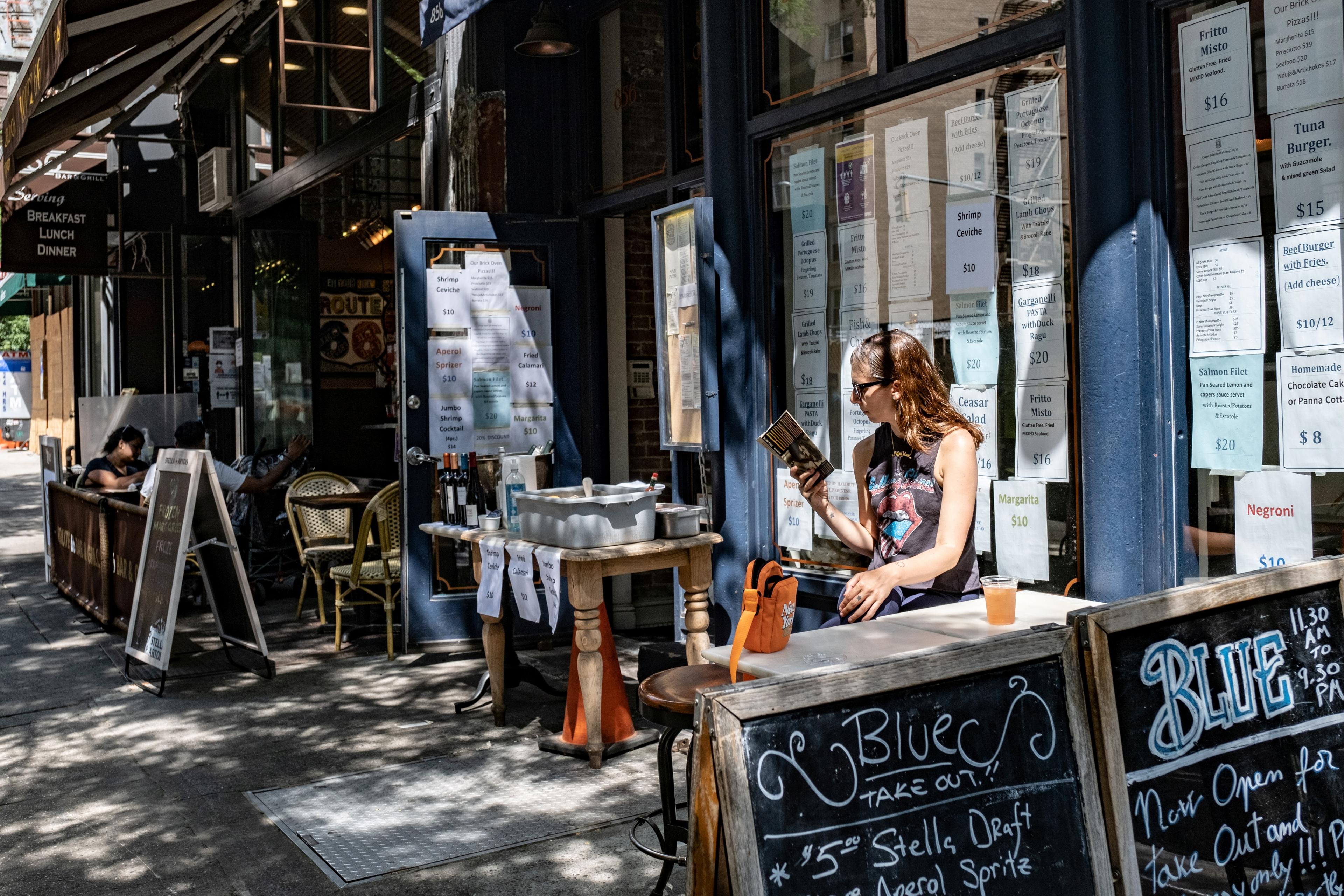 Photo of a woman reading outside a café with menus on windows, chalkboard signs, and outdoor seating on a sunny day.