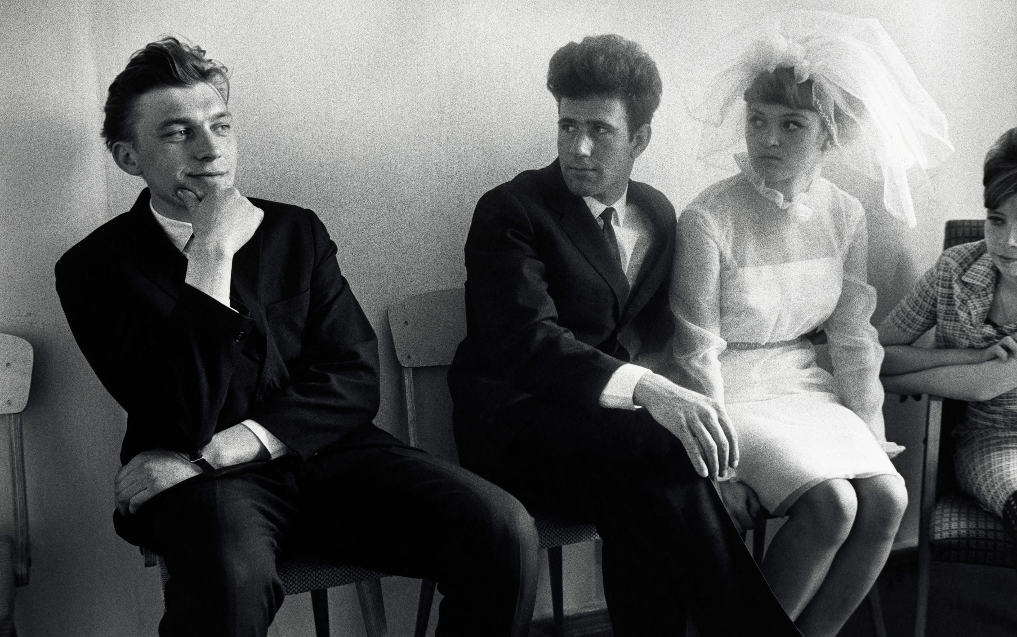 Black-and-white photo of two men in dark suits and a woman in a wedding dress sitting on chairs in a waiting room with blank expressions.