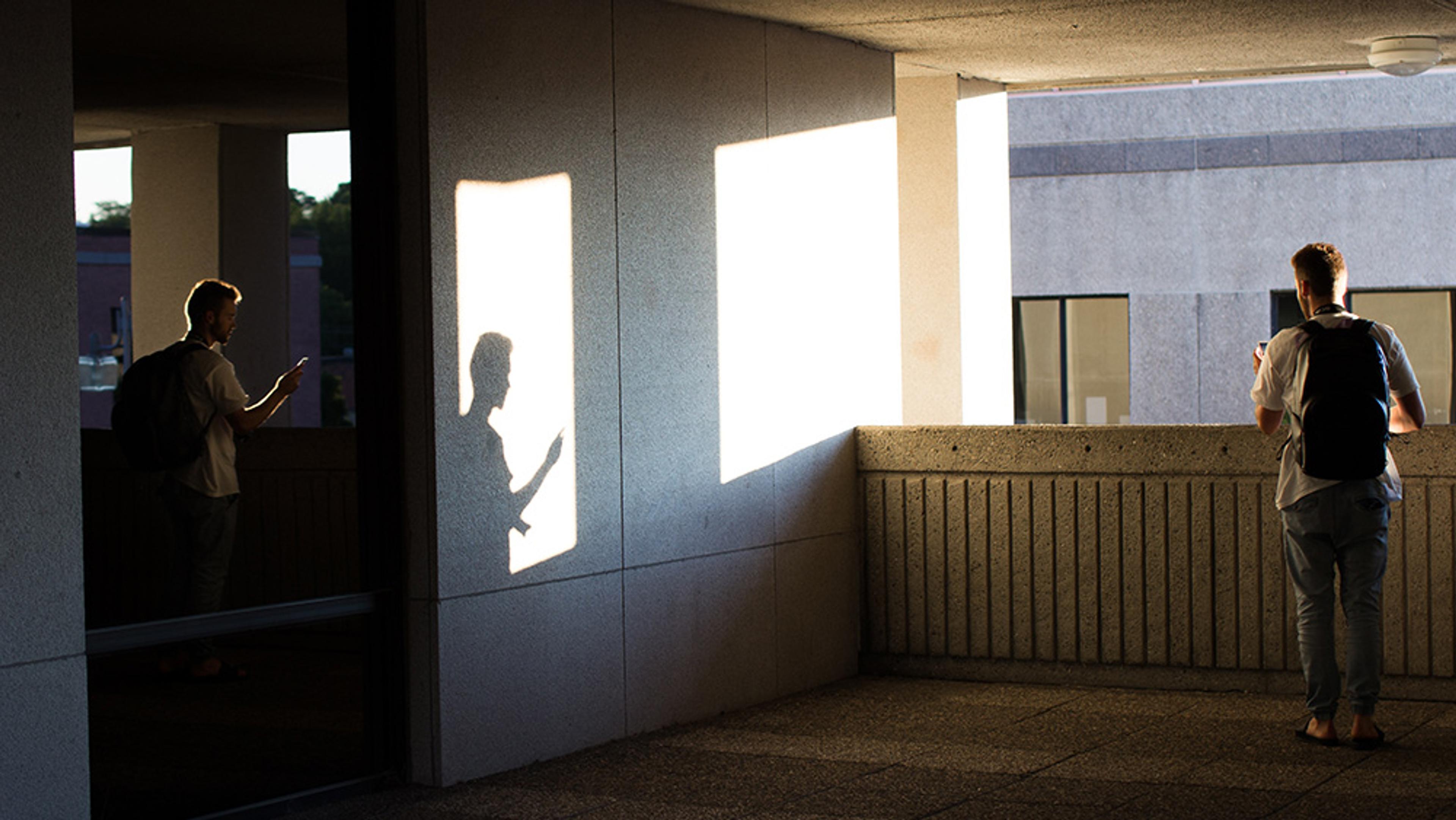 Two men with backpacks standing in a building corridor, using their phones. A shadow of one is cast on a wall.