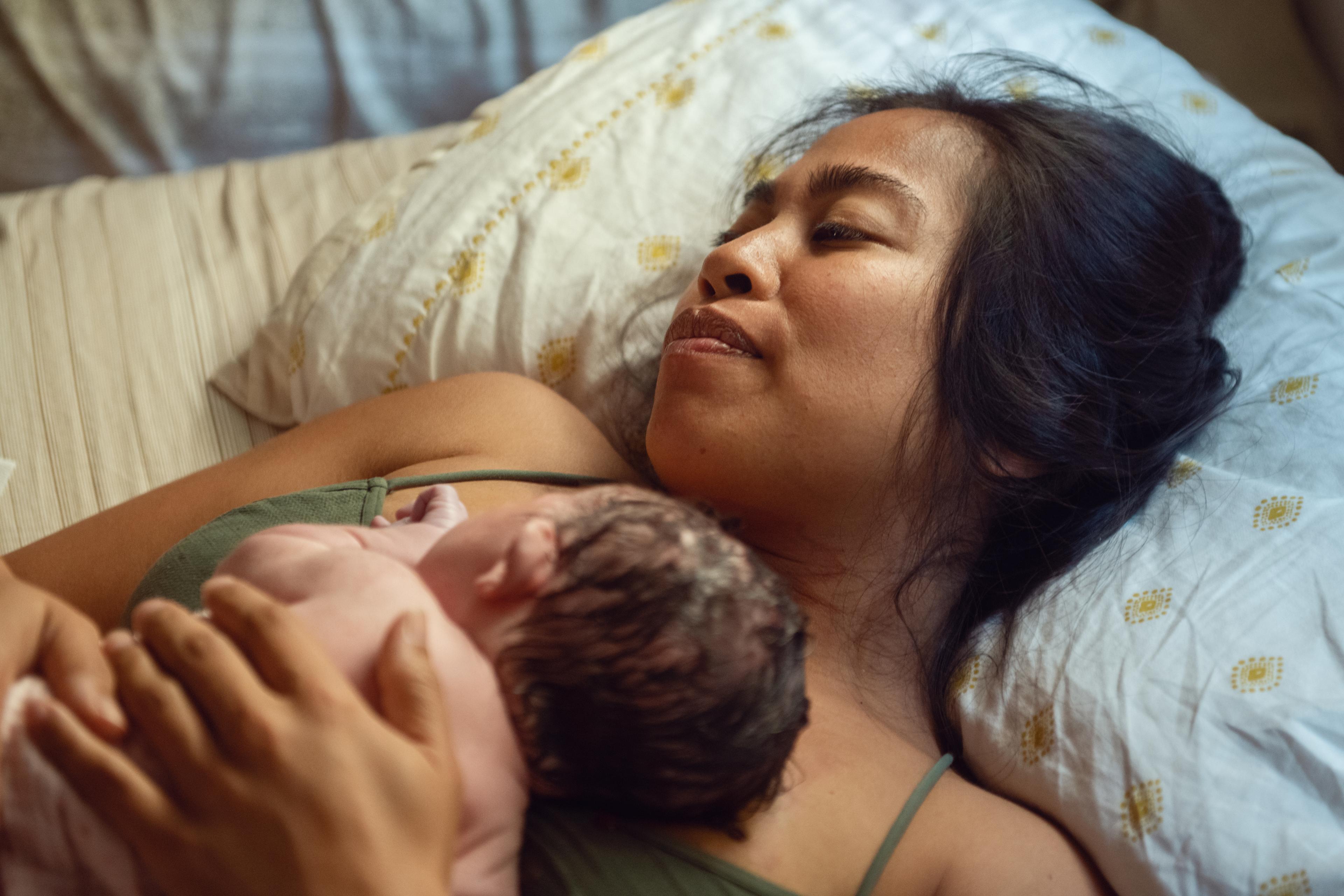 Photo of a woman lying on a bed holding a newborn baby on her chest, both relaxed; soft, warm lighting.
