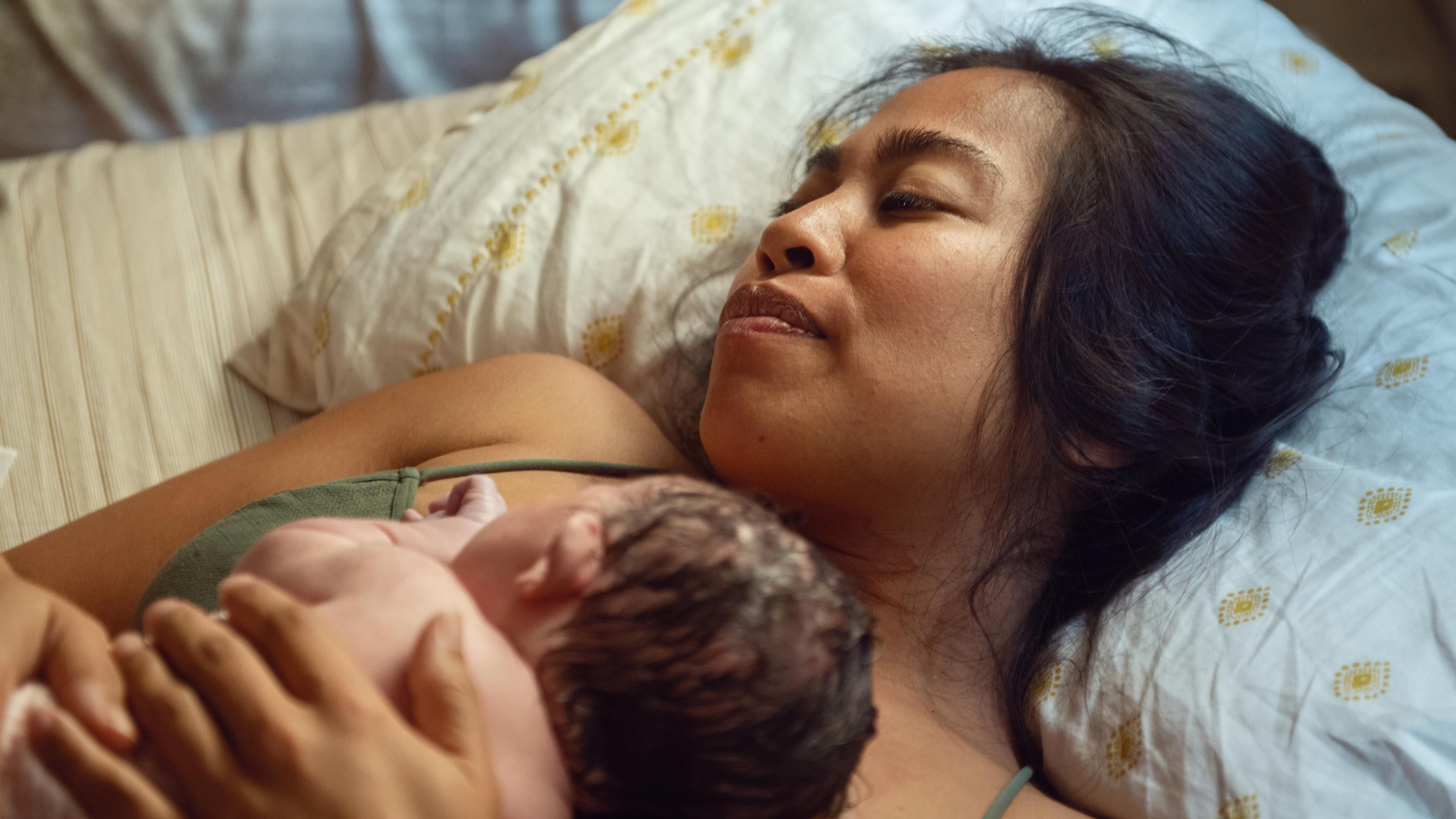 Photo of a woman lying on a bed holding a newborn baby on her chest, both relaxed; soft, warm lighting.