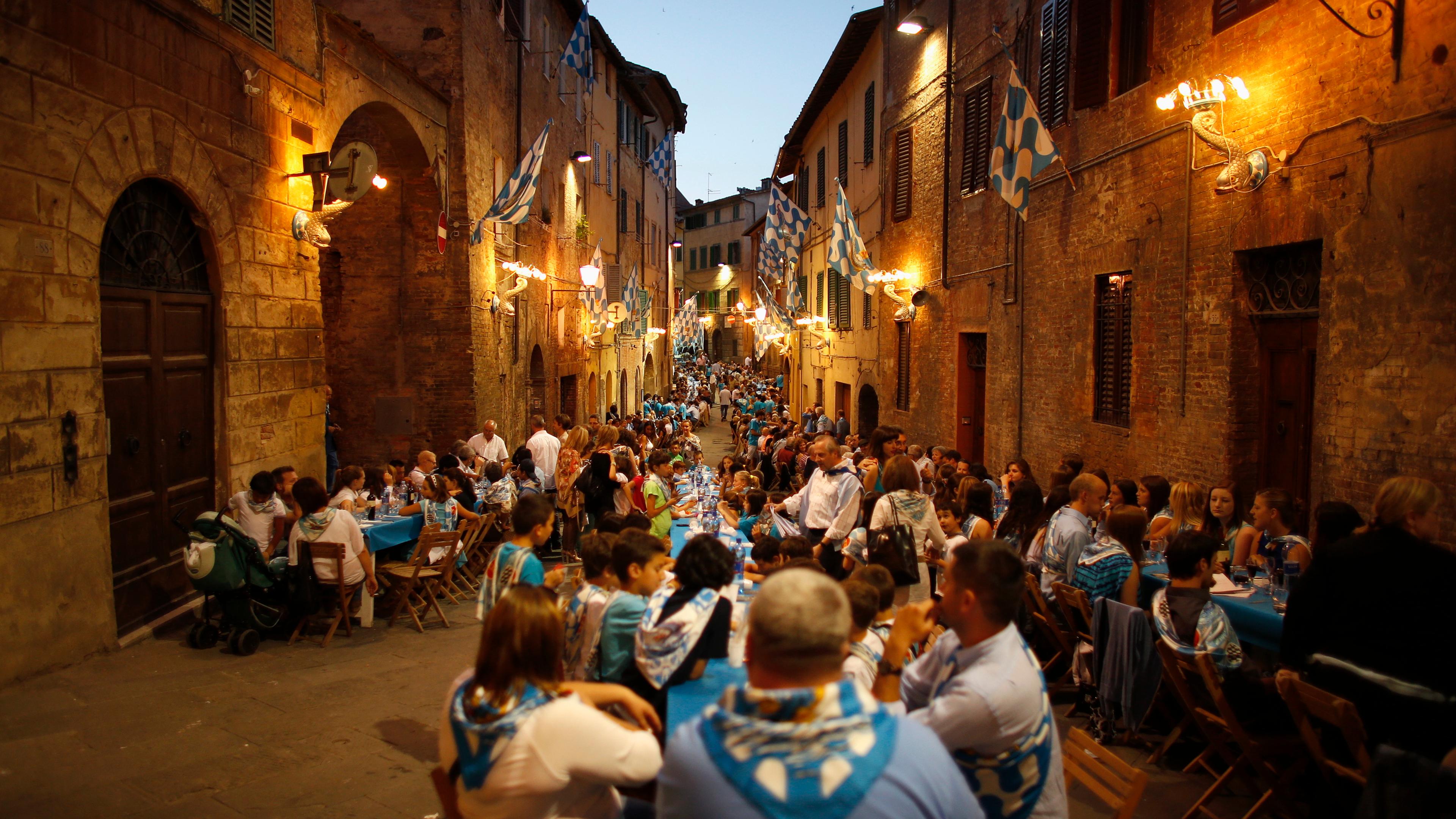 Photo of a crowded street dinner with blue flags, people sitting at long tables amid warm lights in a historic setting.