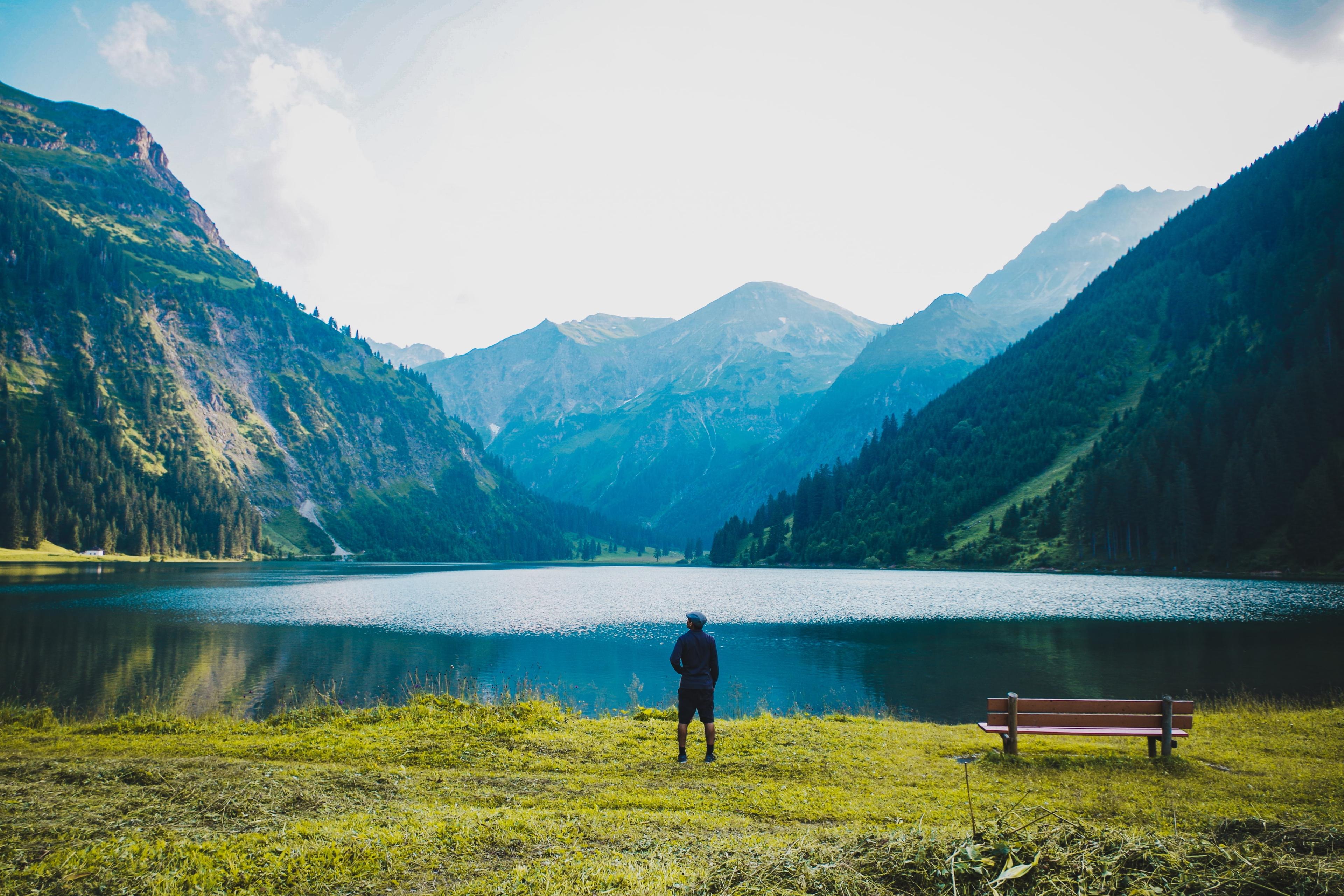 Photo of a person standing by a mountain lake with a bench nearby, surrounded by lush green mountains under a clear sky.