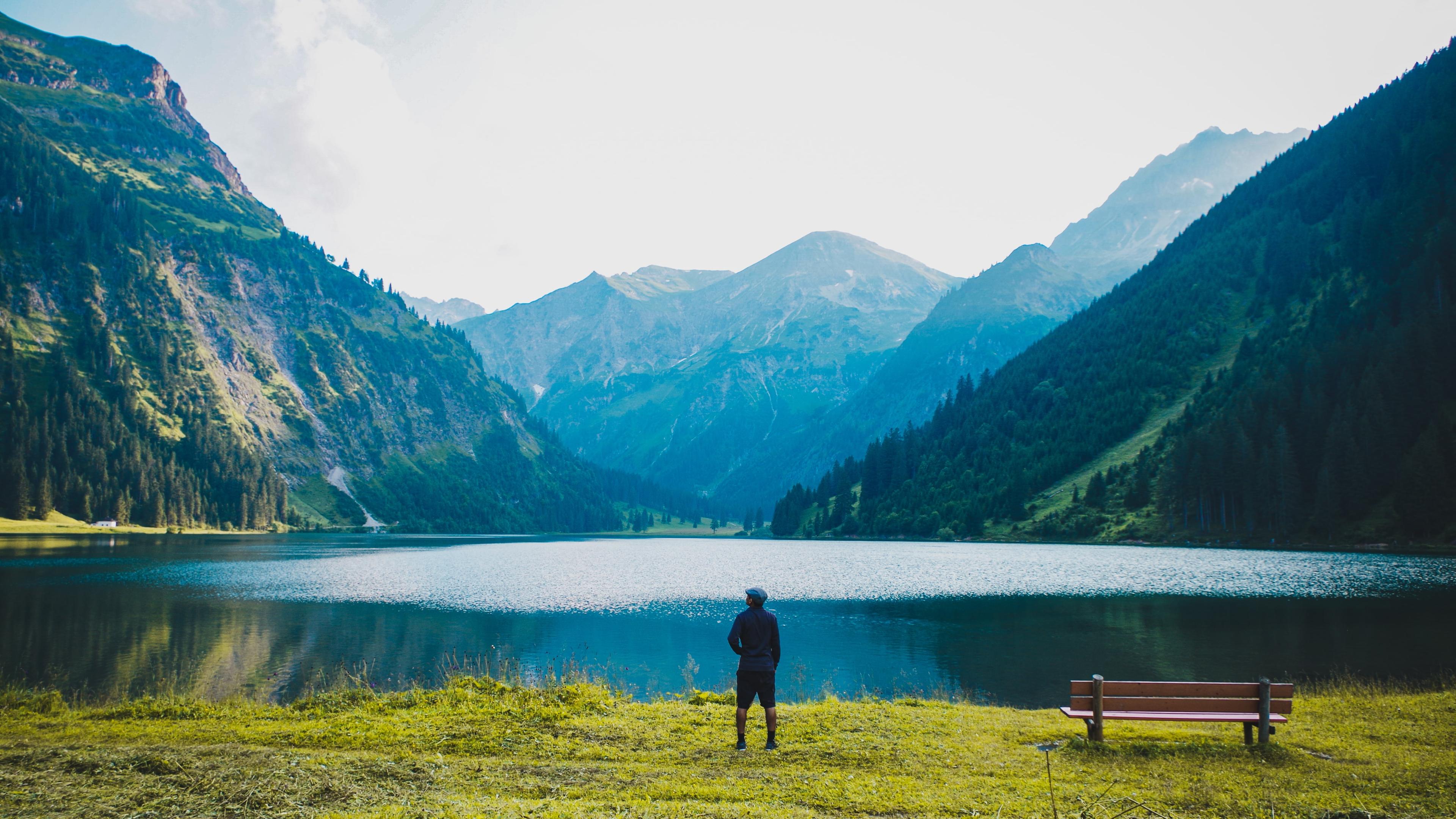 Photo of a person standing by a mountain lake with a bench nearby, surrounded by lush green mountains under a clear sky.