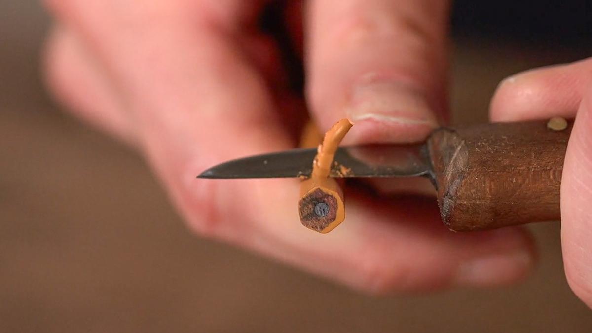 Close-up photo of a hand carving a pencil with a small knife, focusing on the pencil’s tip and wood shavings.