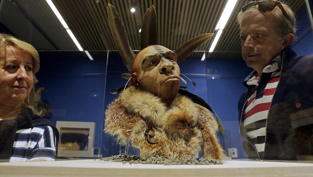 Two people look at a scientifically based recreation of a Neanderthal man’s head, adorned with feathers and fur, enclosed in a glass case at a museum. The setting is under strong indoor lighting with a blue wall background.