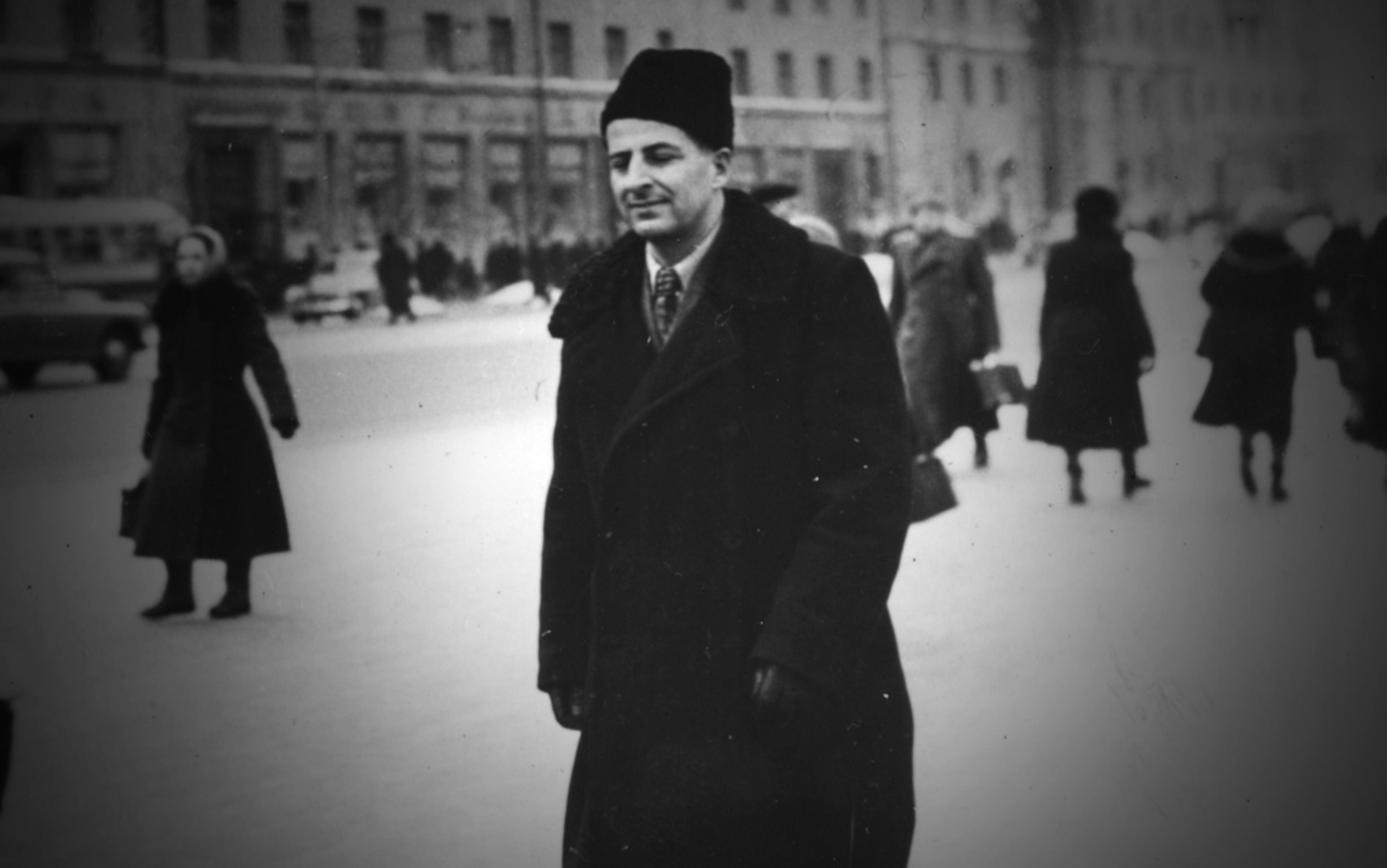 Black-and-white photo of a man in a winter coat and hat walking on a city street with people in the background, mid-20th century.