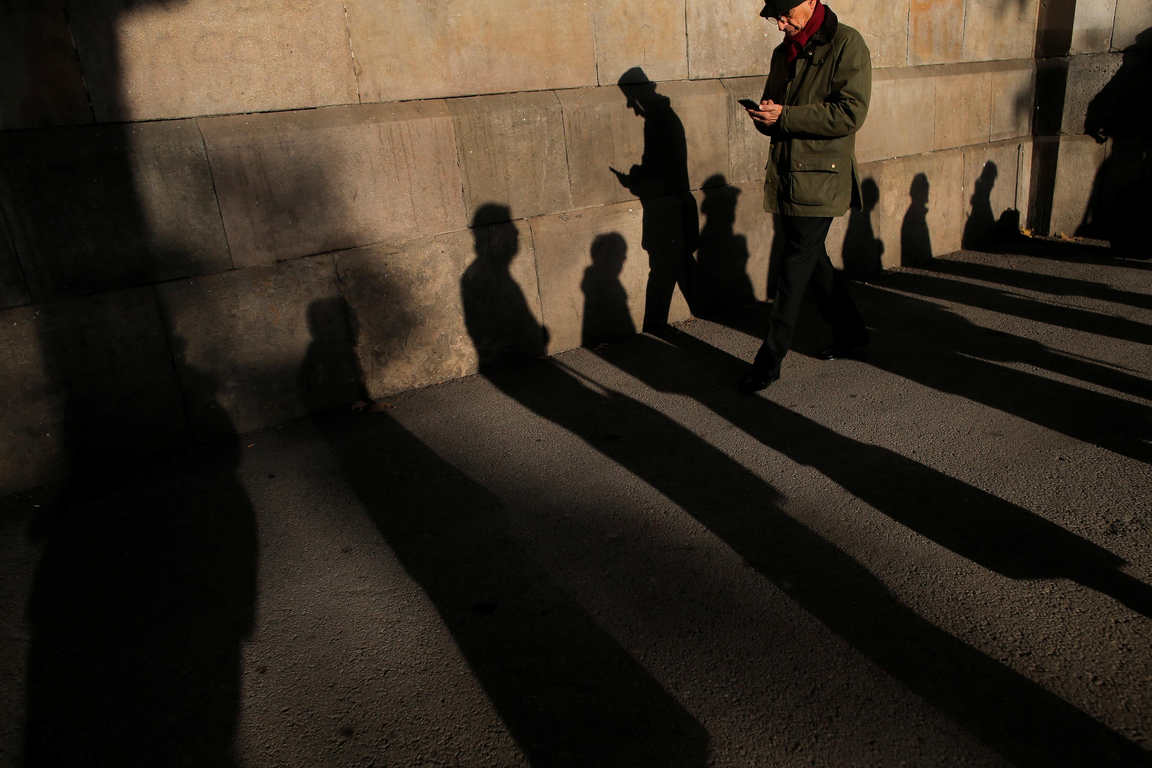Photo of a person walking, casting long shadows on a paved ground near a wall. Shadows of other unseen people are also visible.