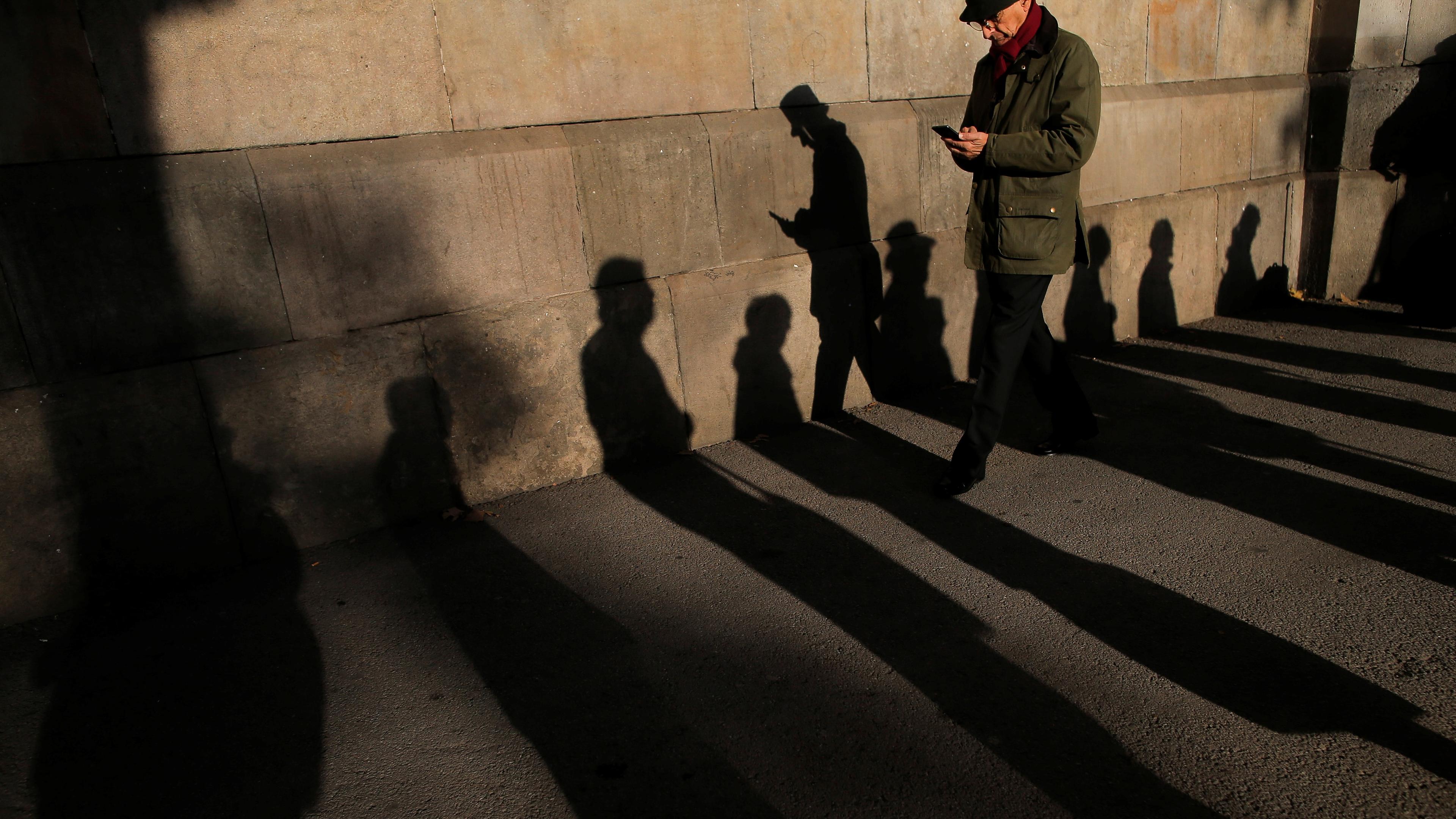 Photo of a person walking, casting long shadows on a paved ground near a wall. Shadows of other unseen people are also visible.
