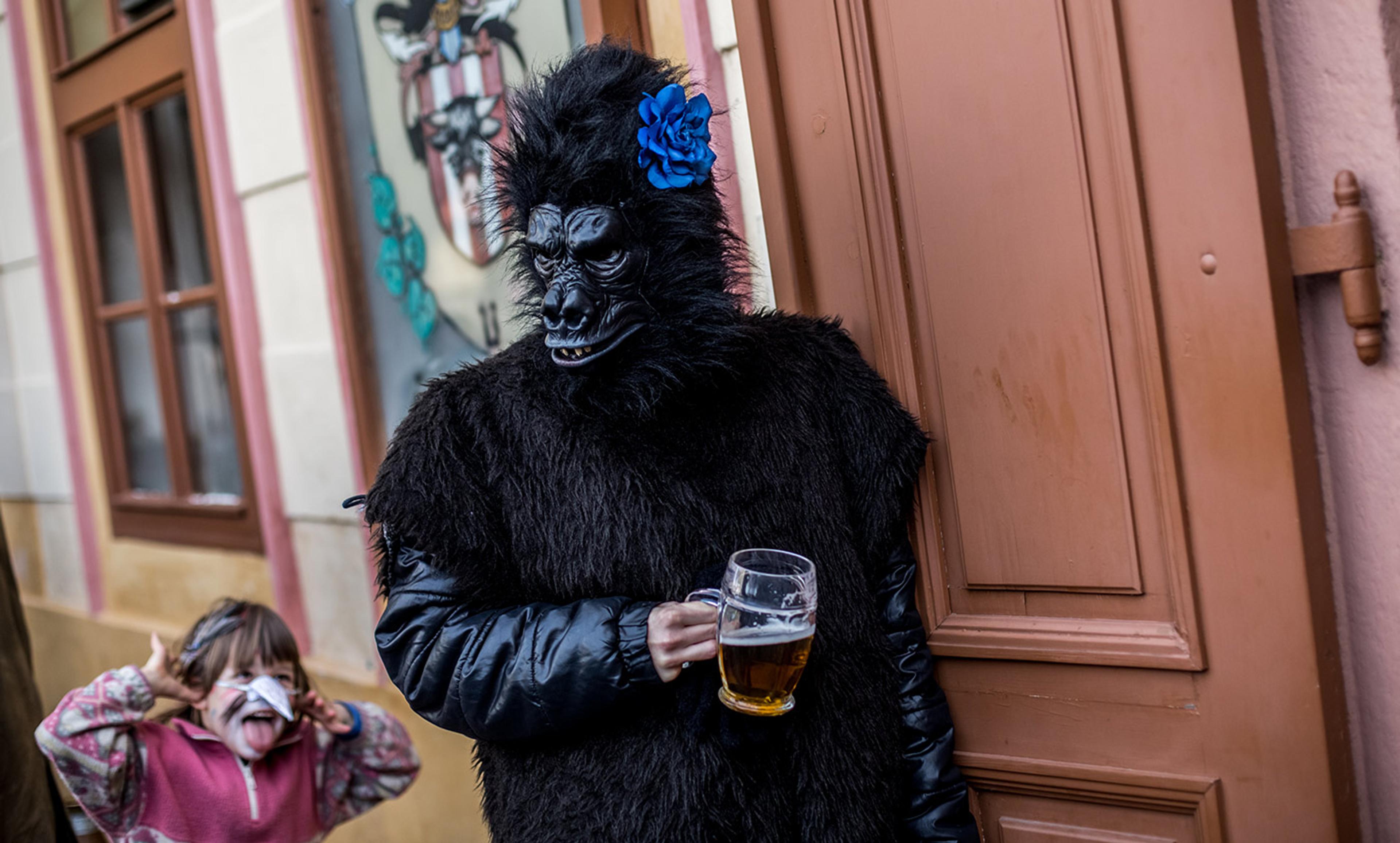 A person in a gorilla costume holding a beer, with a child making a face in the background, standing in front of a building.