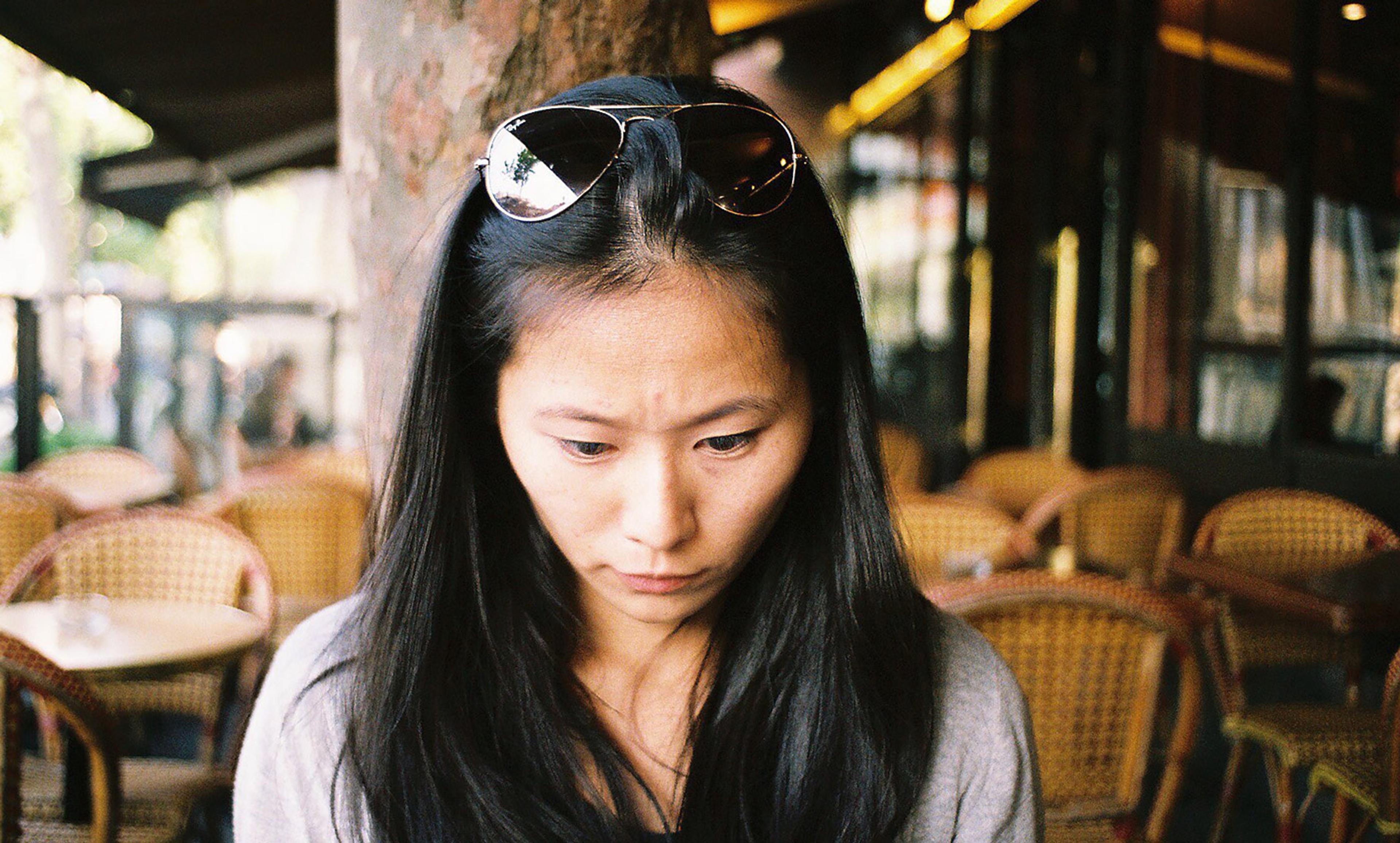 Asian woman with dark hair and sunglasses on her head, looking down at a table in an outdoor café with wicker chairs.